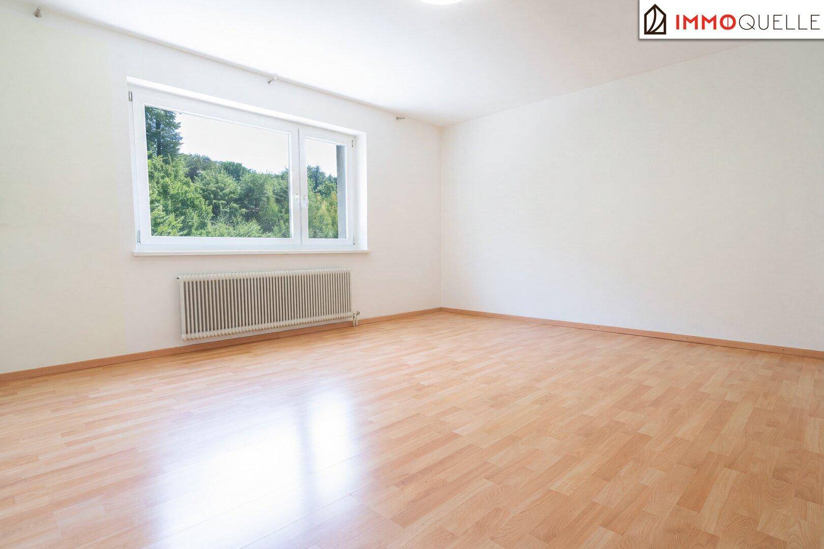 Helles, leeres Schlafzimmer mit Holzboden und großem Fenster mit Blick auf Bäume.
