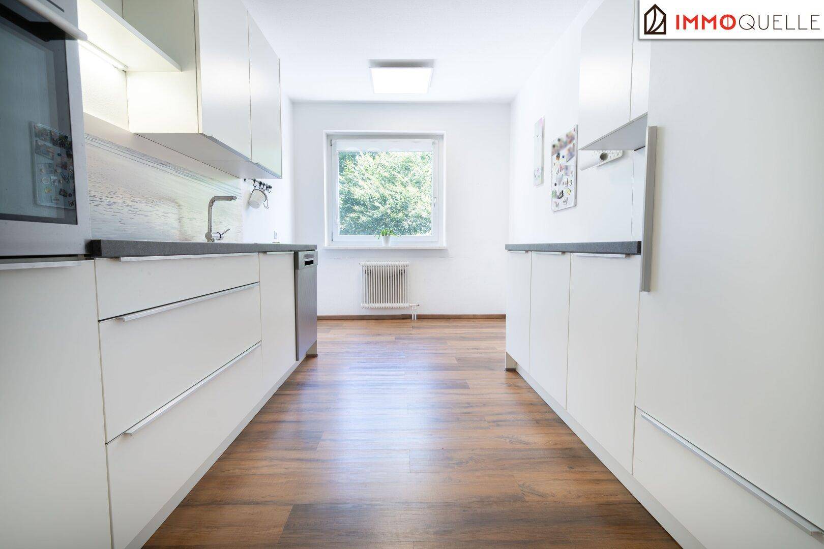 Moderne Küche mit weißen Schränken, dunklen Arbeitsplatten und Fenster mit Grünblick.