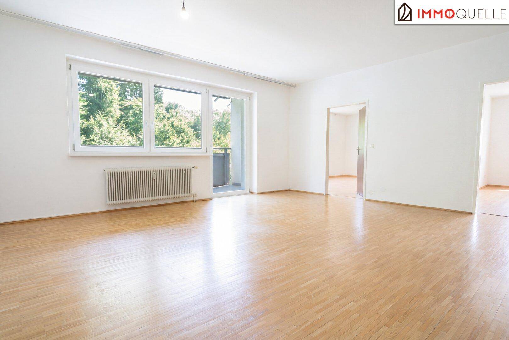 Geräumiges, leeres Wohnzimmer mit Holzboden, großem Fenster und Zugang zum Balkon.