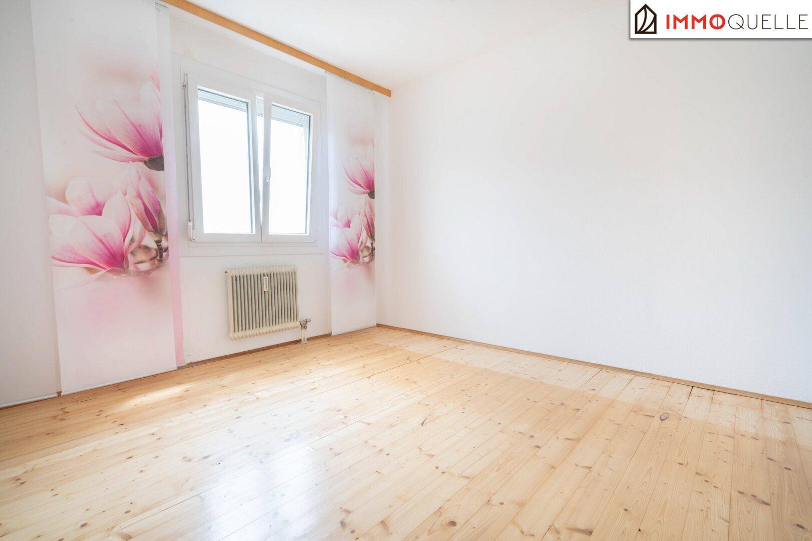 Heller Raum mit Holzfußboden, großem Fenster und floralen Dekorationselementen an den Wänden.