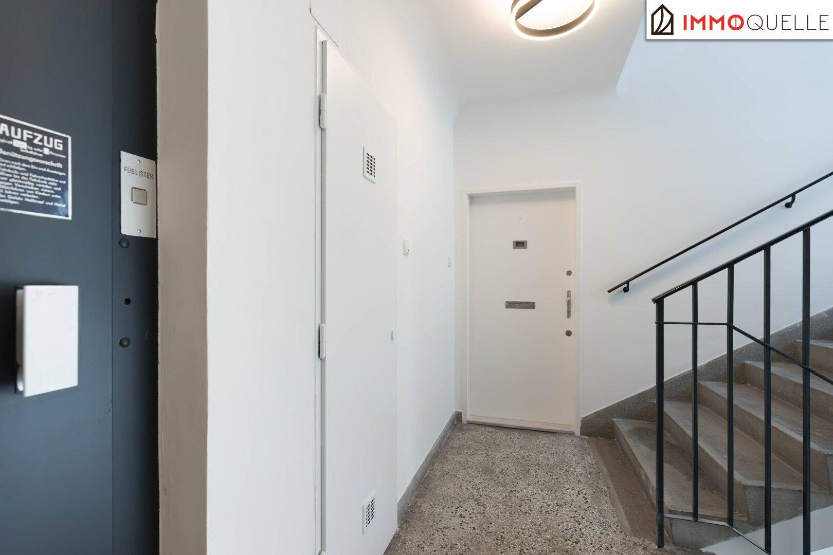Heller Hausflur mit Aufzug, weißer Wohnungstür und Treppenaufgang mit schwarzem Geländer.