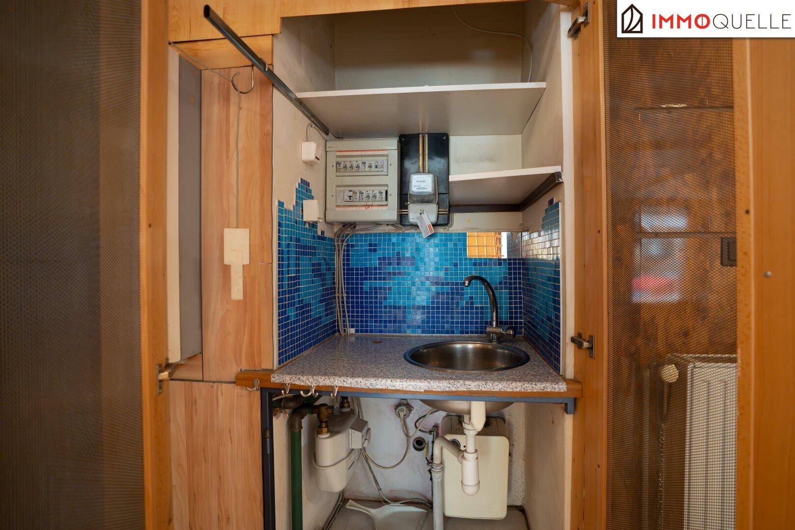 Kleine, eingebaute Küchenzeile mit Spülbecken, blauen Mosaikfliesen und elektrischen Installationen in einem Schrank.