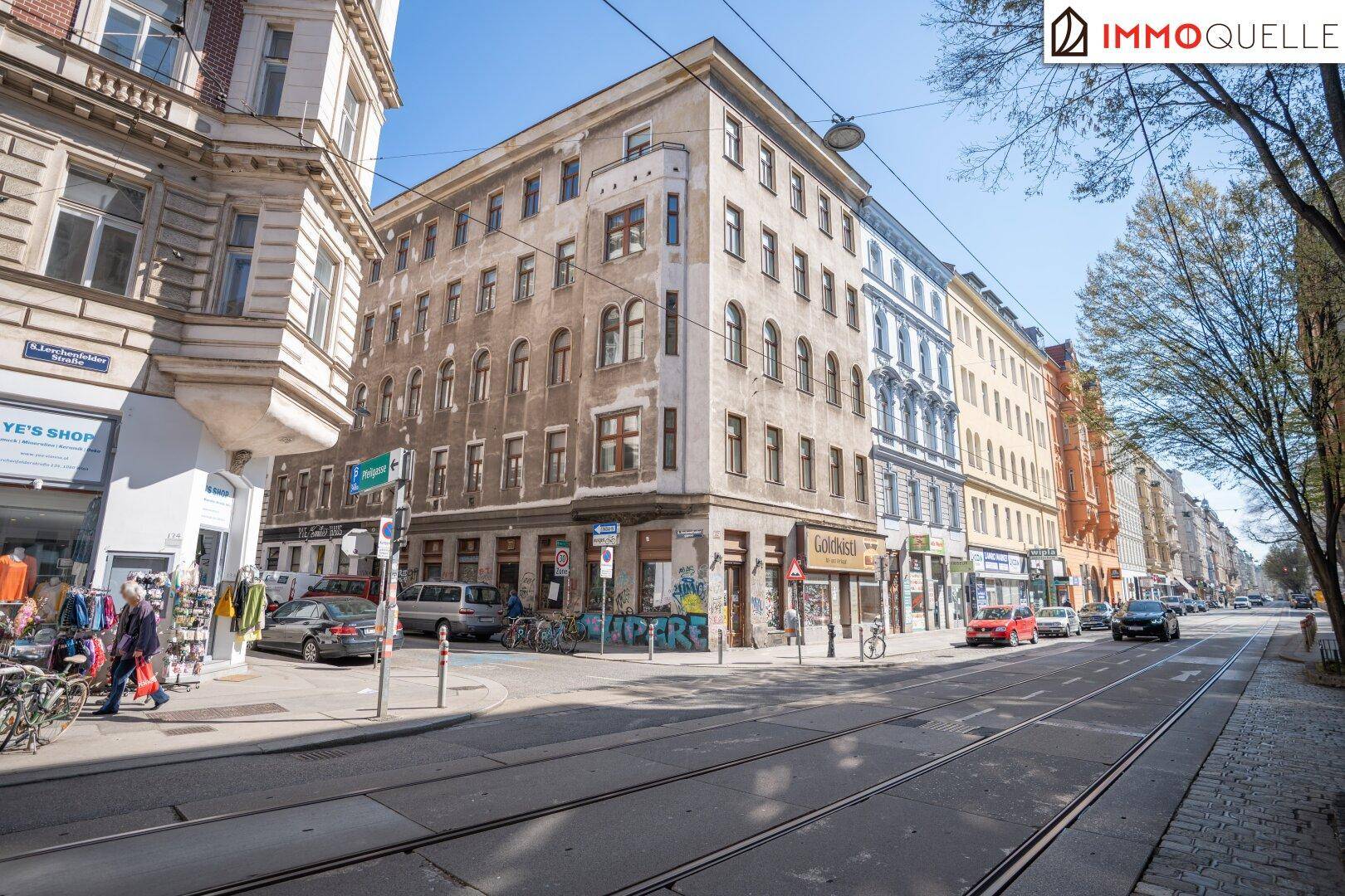 Weitläufiger Straßenblick mit Eckgebäude, geparkten Autos und Passanten an einem sonnigen Tag.