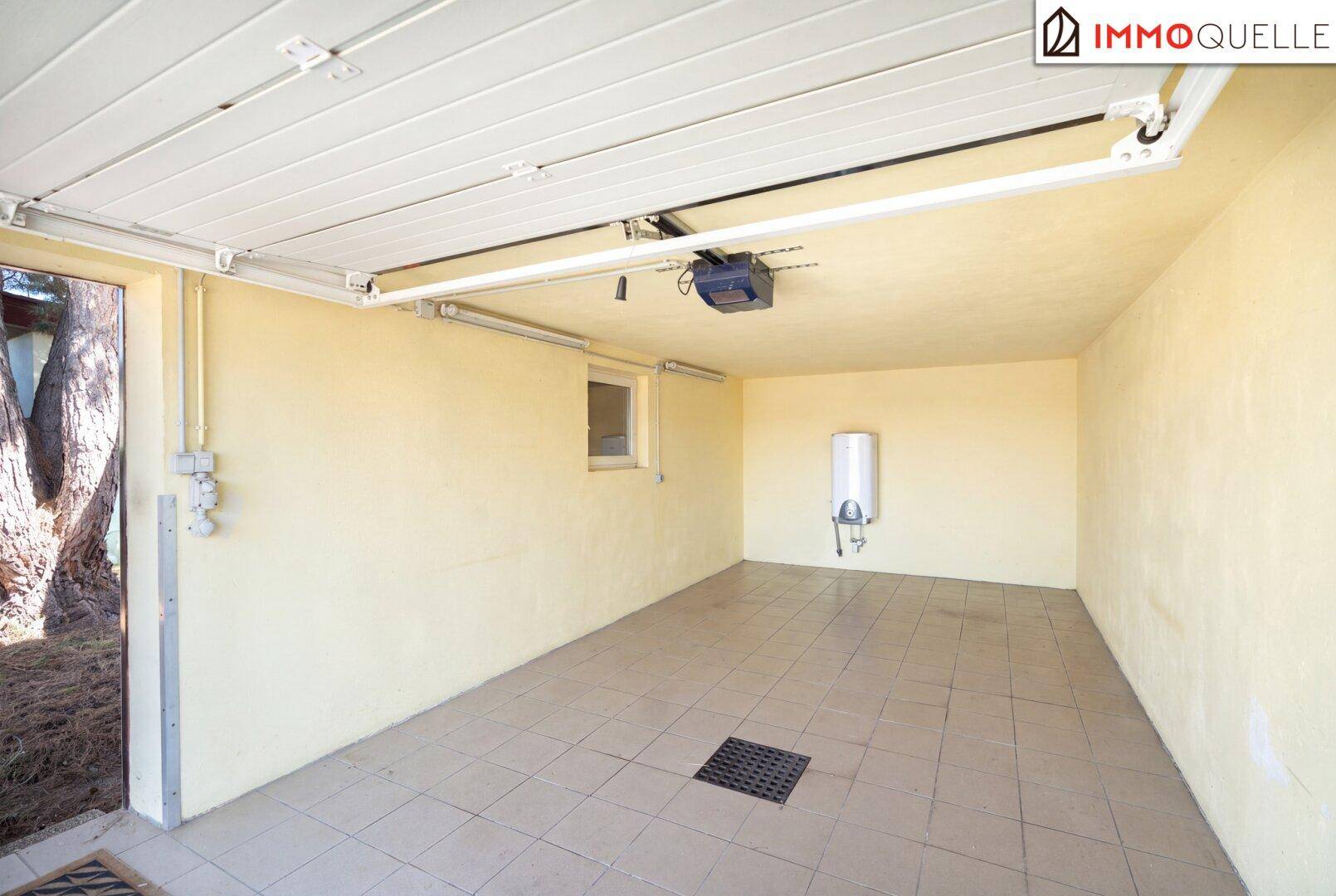 Geräumige Garage mit Fliesenboden, Fenster und einem Warmwasserboiler an der Wand.