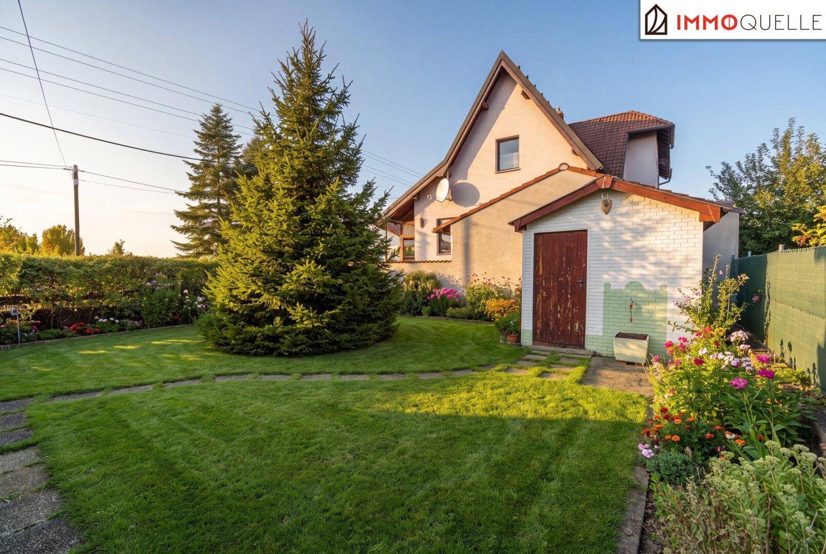 Rückansicht des Hauses mit großem, gepflegtem Garten, Blumenbeeten und einem kleinen Gartenhaus.