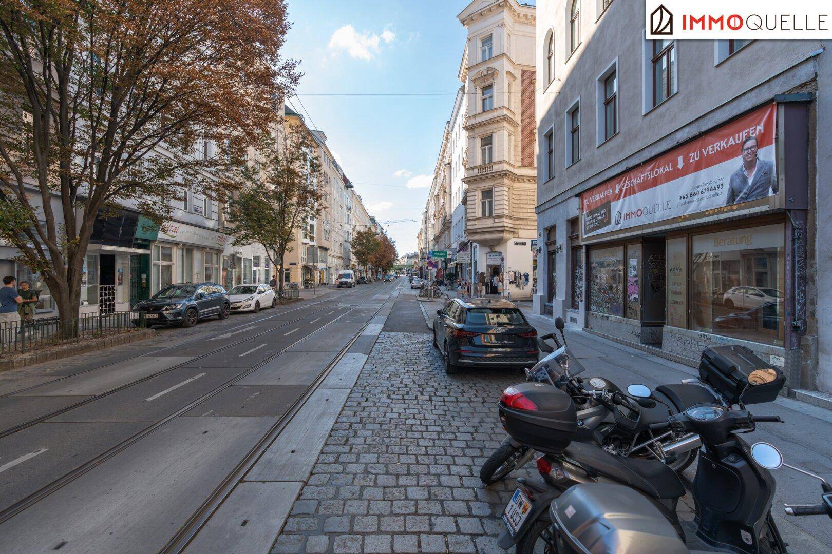 Blick auf eine belebte Stadtstraße mit historischen Gebäuden, geparkten Fahrzeugen und Straßenbahnschienen.