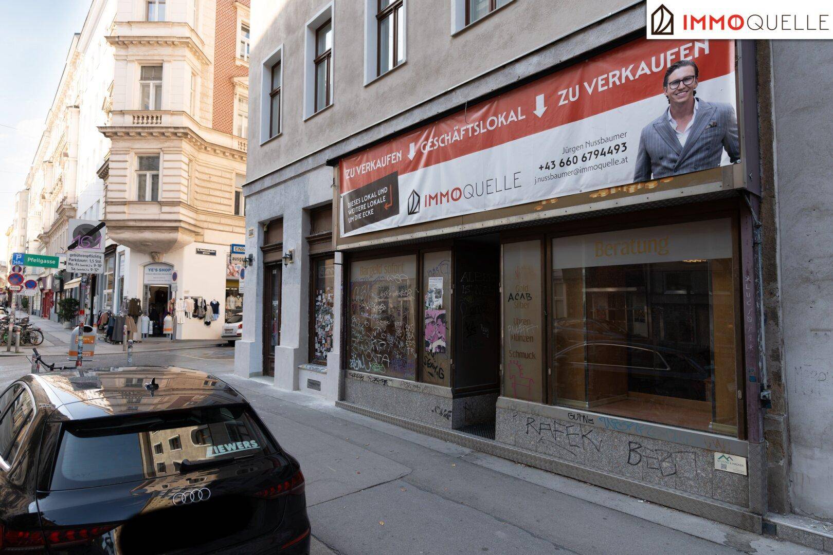 Gebäudefassade mit Geschäftsräumen im Erdgeschoss und einem 'Zu Verkaufen'-Banner. Ein schwarzes Auto parkt auf der Straße.