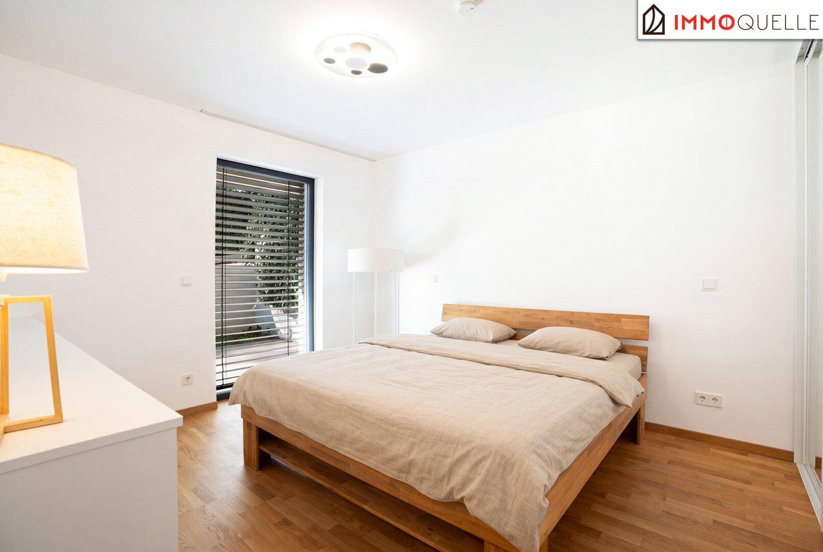 Helles Schlafzimmer mit Holzbett, Parkettboden und großem Fenster mit Jalousien.