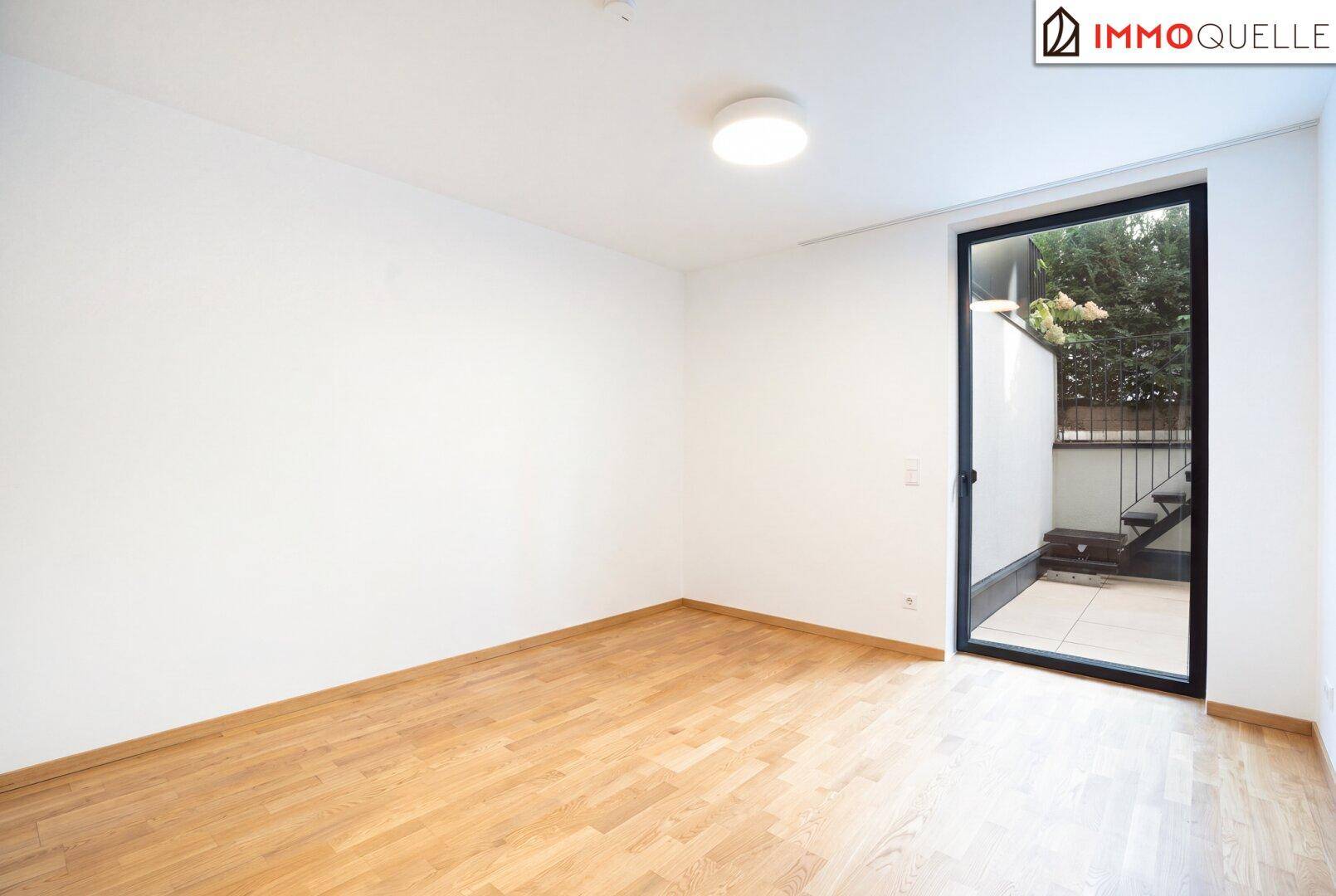Leeres Zimmer mit Holzboden und großem Fenster, das Zugang zu einem Außenbereich bietet.