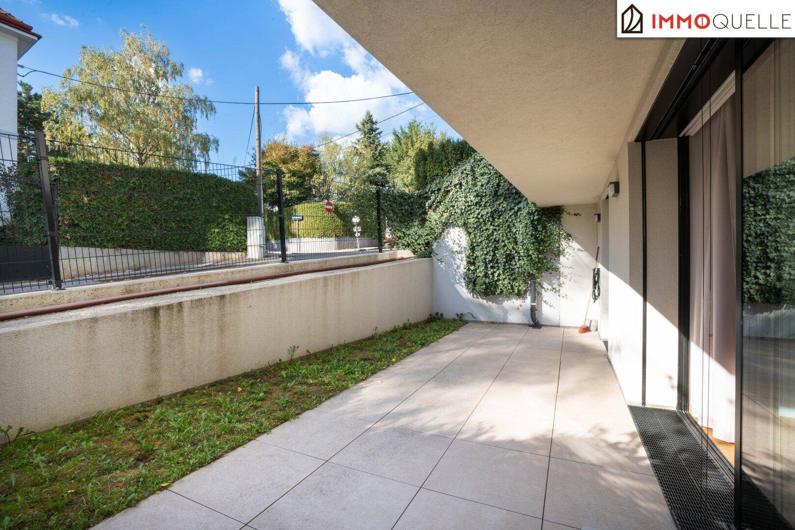 Unmöblierte Terrasse mit Fliesenboden und kleiner Rasenfläche, umgeben von einer Hecke und Mauer.