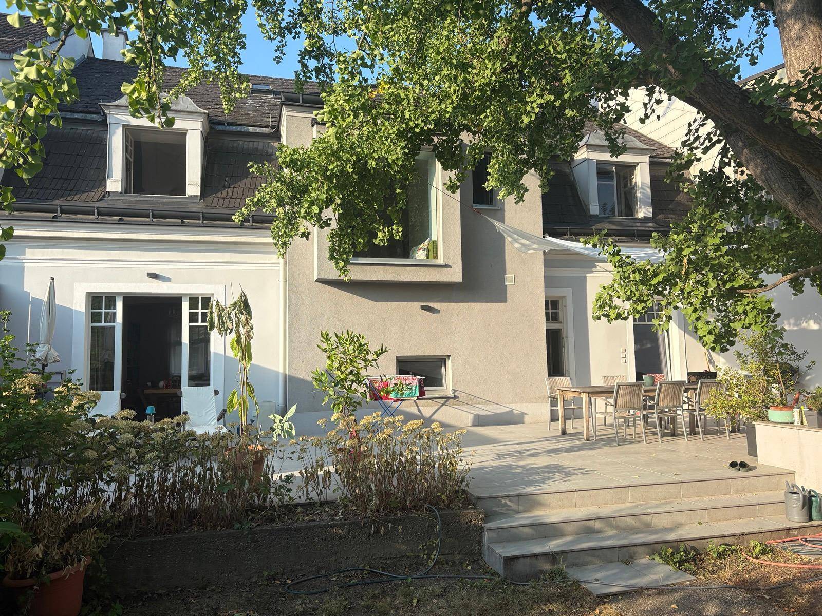 Hinteransicht des Hauses mit großer Terrasse, Gartenmöbeln und üppiger Bepflanzung.