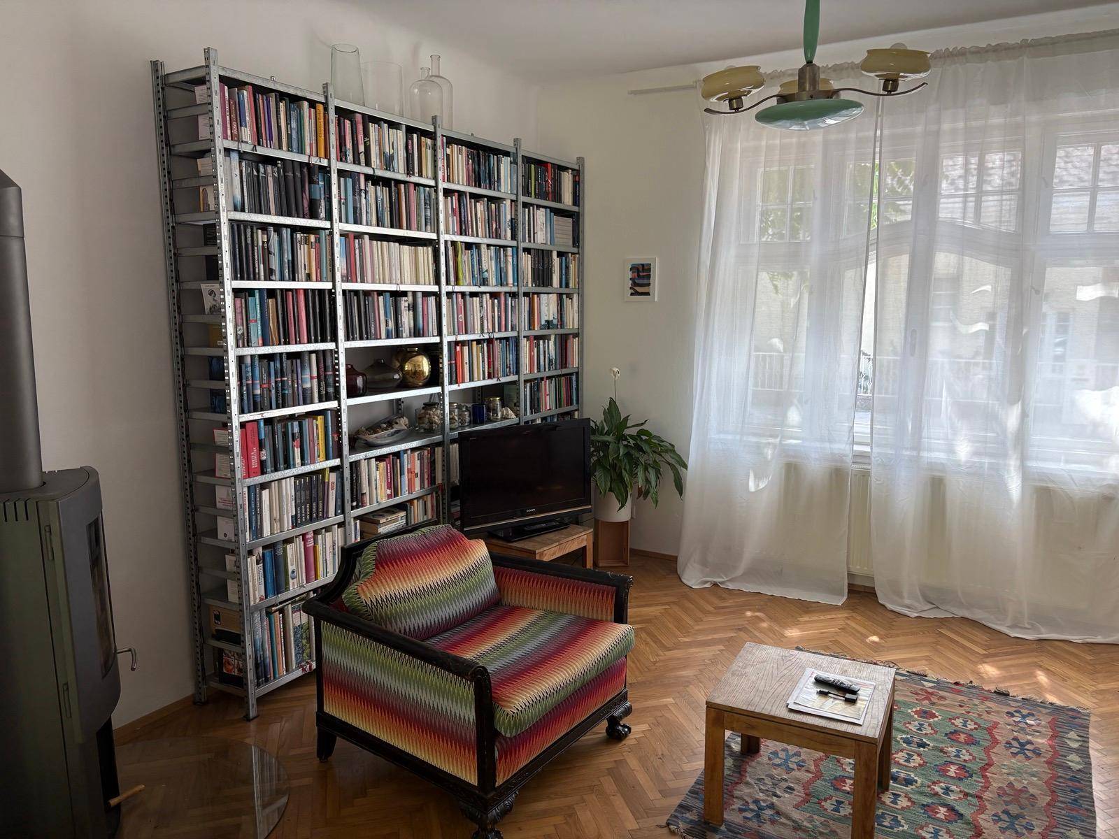 Gemütliches Wohnzimmer mit Bücherregal, Sessel und Parkettboden, hell durch große Fenster.