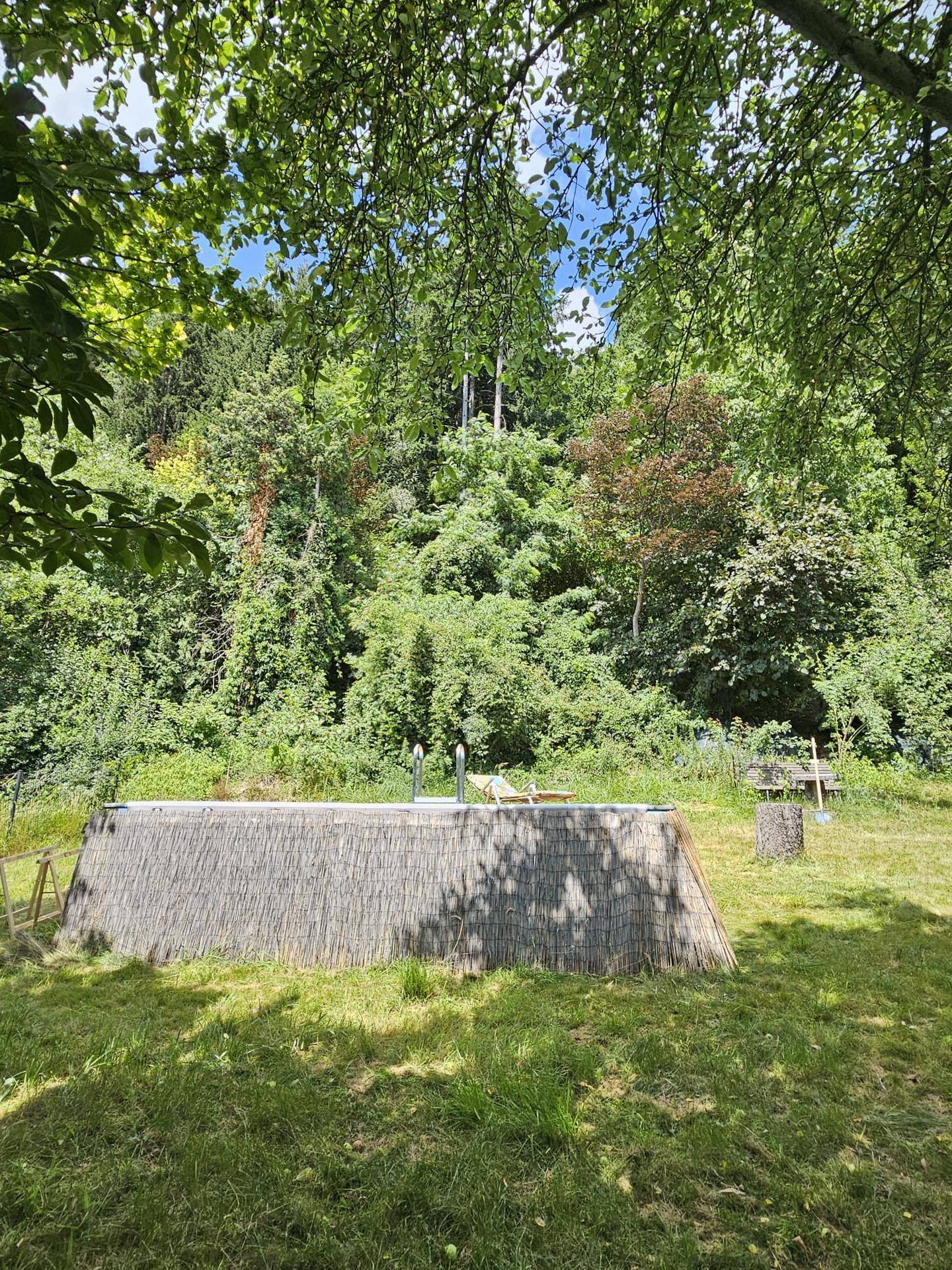 Gartenbereich mit einem umzäunten Aufstellpool, umgeben von üppiger Vegetation und Bäumen.