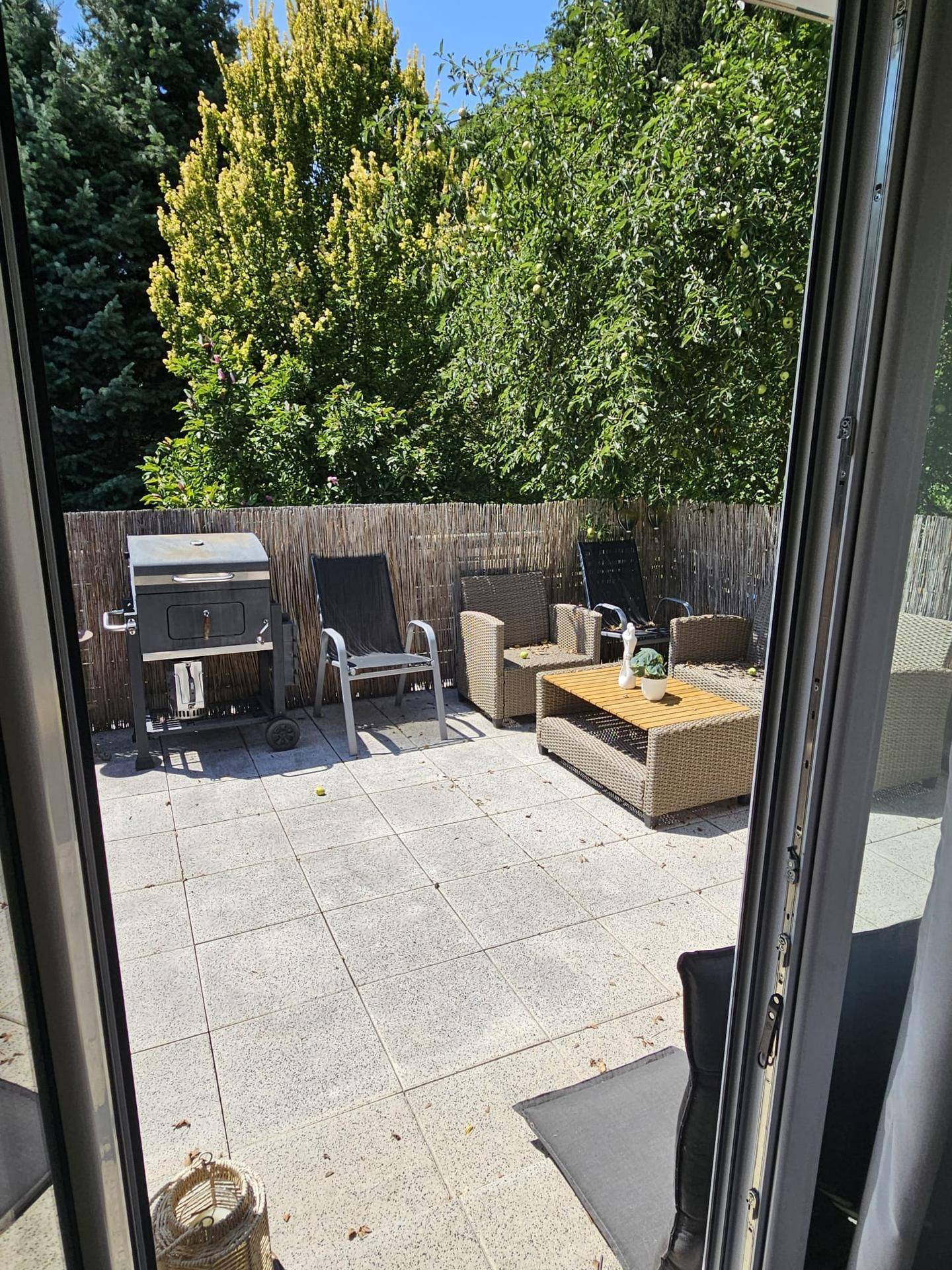 Geflieste Terrasse mit Grillbereich und Gartenmöbeln, umgeben von Grün und Zaun.