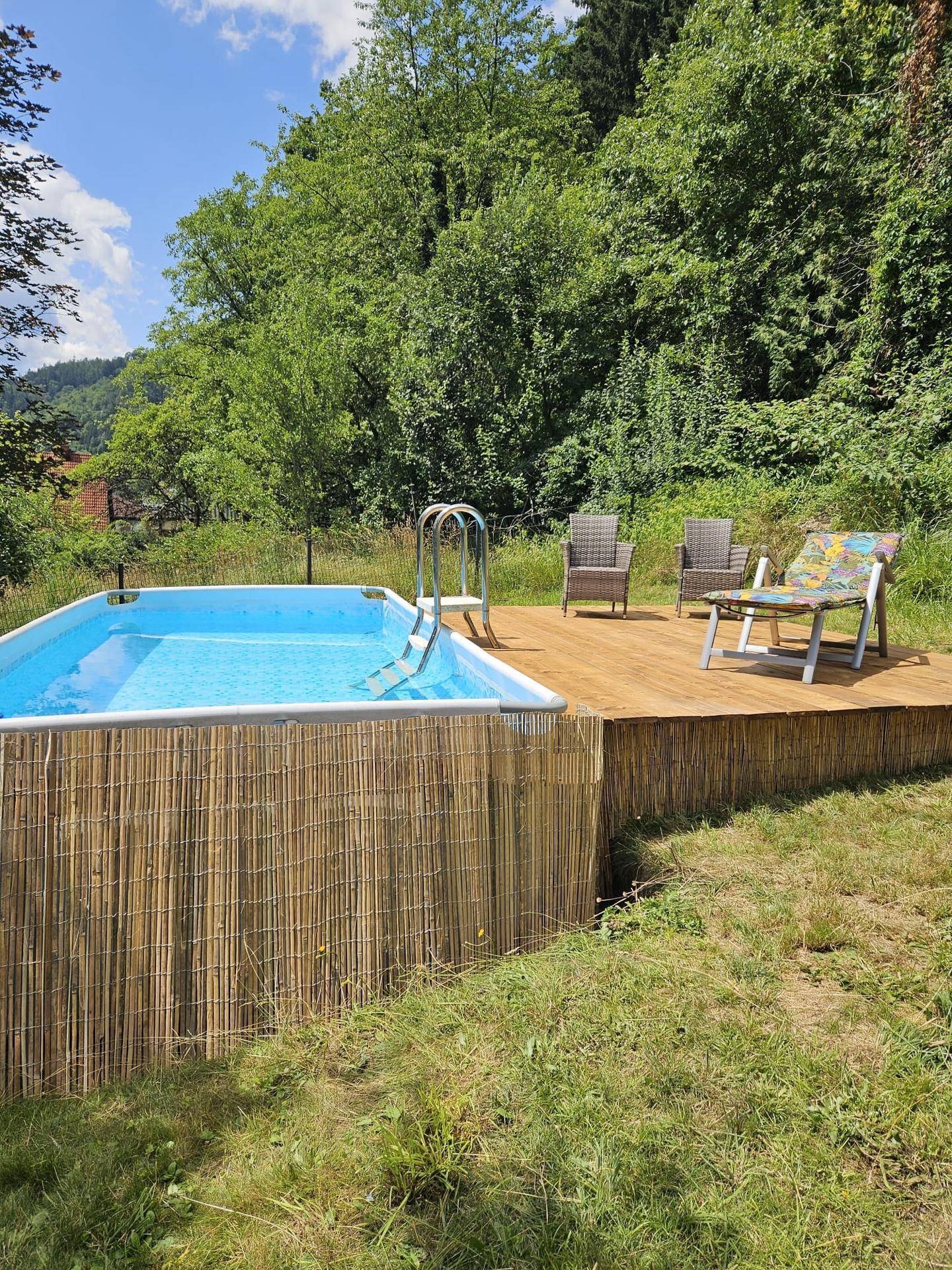 Aufstellpool mit Holzdeck und Liegestühlen, umgeben von viel Grün unter blauem Himmel.