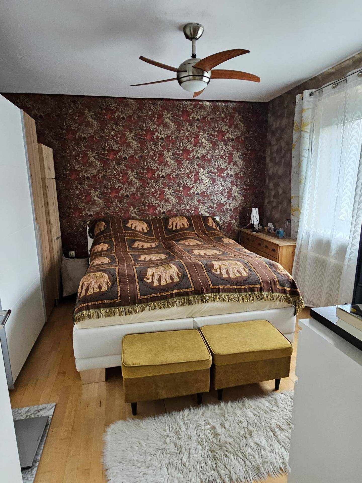 Schlafzimmer mit Doppelbett, Deckenventilator und gemusterter Tapete an der Wand.