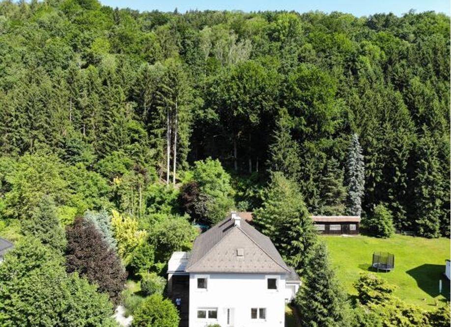 Luftaufnahme des Hauses umgeben von dichtem Wald und weitläufiger Grünfläche mit Trampolin.