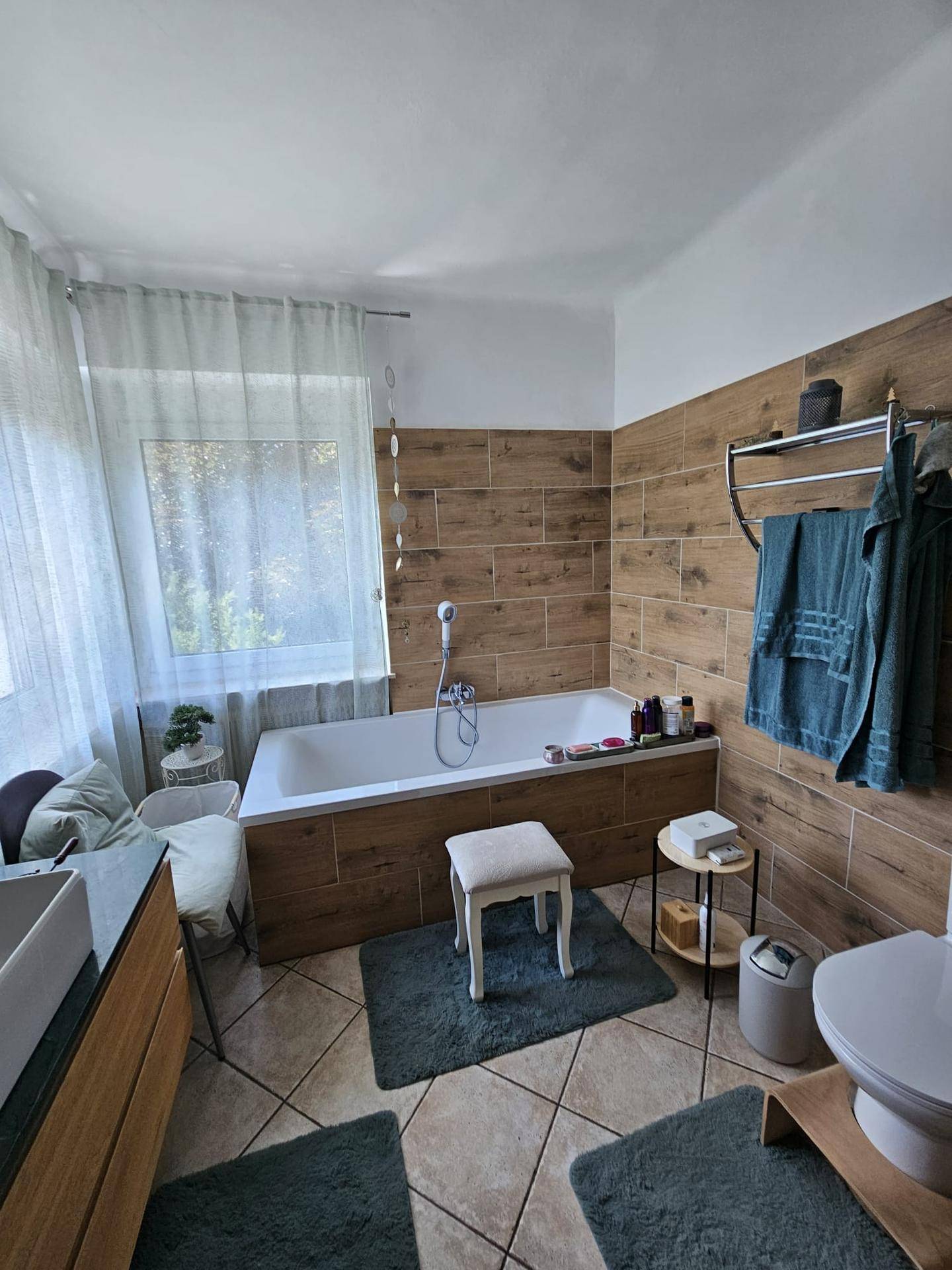 Badezimmer mit Badewanne, Holzoptik-Fliesen und Fenster für natürliches Licht.