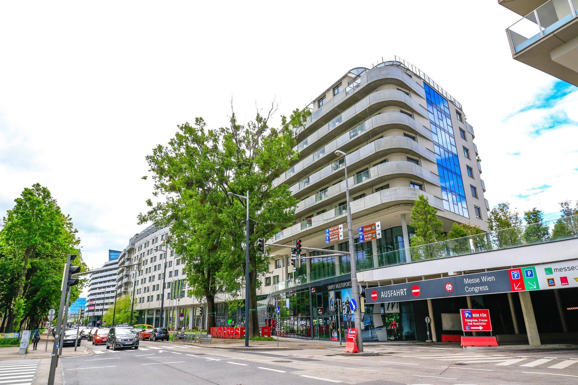 Modernes Gebäude an einer belebten Straße mit Geschäften im Erdgeschoss und Tiefgaragenzufahrt.