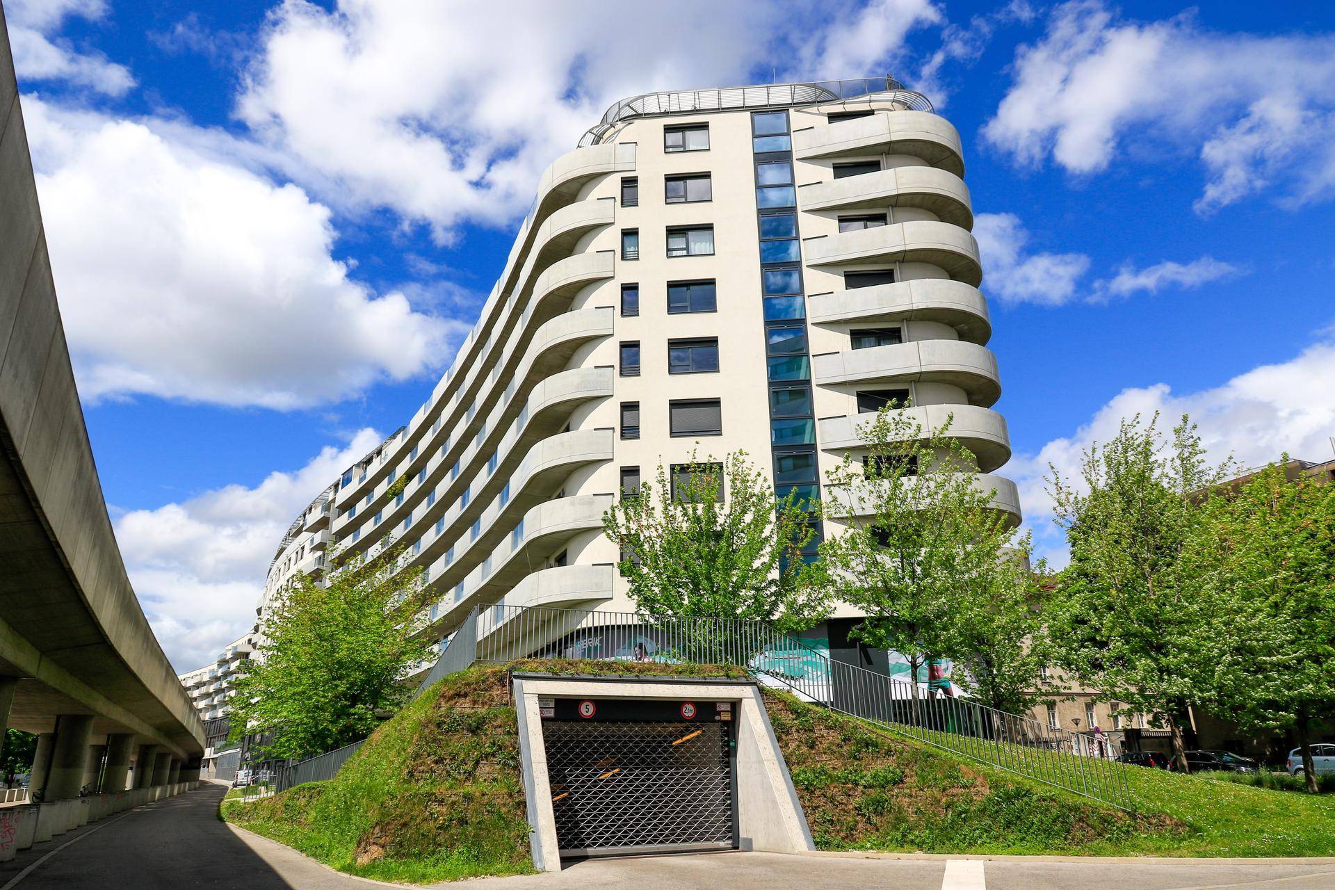 Modernes Wohngebäude mit geschwungenen Balkonen und Tiefgaragenzufahrt unter blauem Himmel.