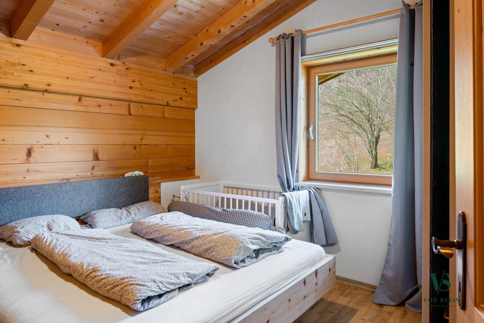 Helles Schlafzimmer mit Holzwänden, Holzboden und einem Fenster mit Blick ins Grüne.