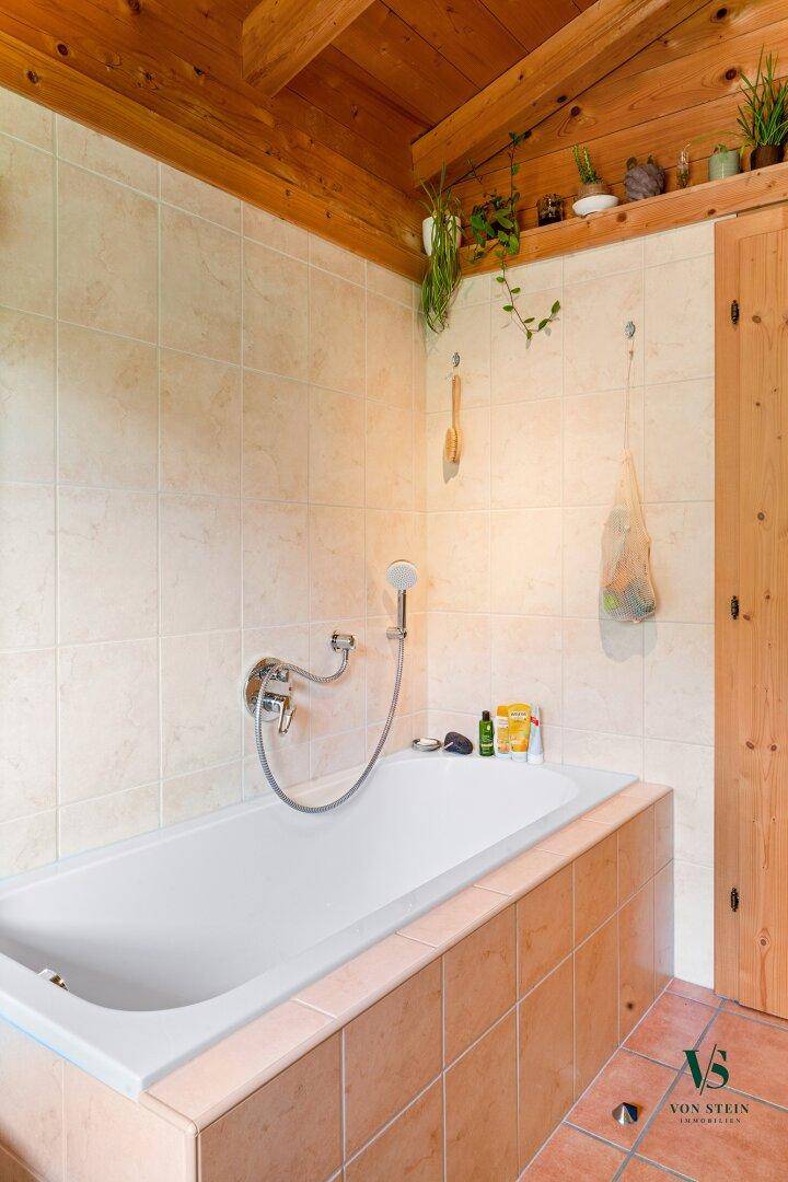 Badezimmer mit Badewanne, beigefarbenen Fliesen und Holzbalkendecke im Landhausstil.
