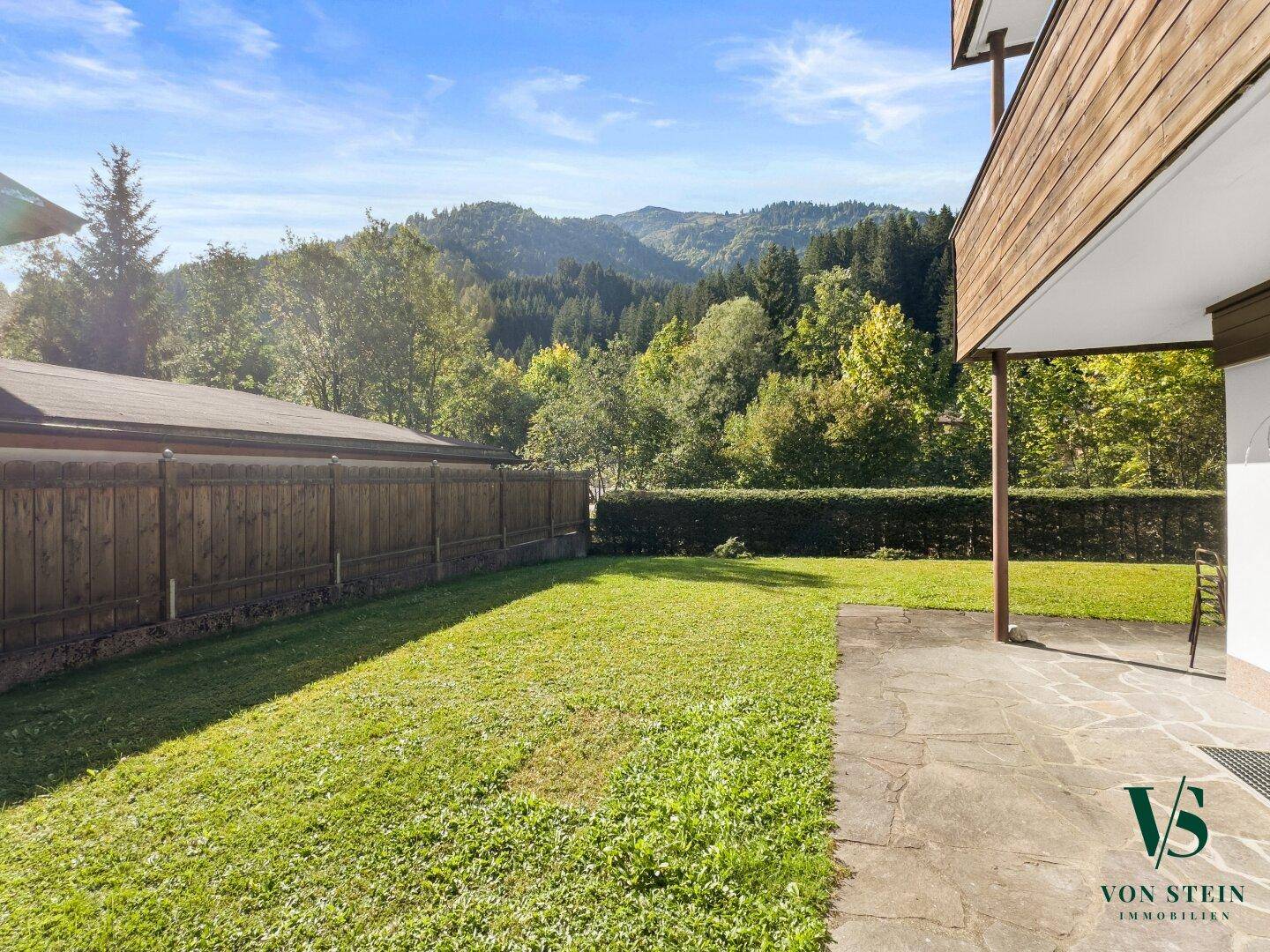 Gepflegter Gartenbereich mit Rasenfläche und Steinterrasse, umgeben von grünen Wäldern und Bergen.