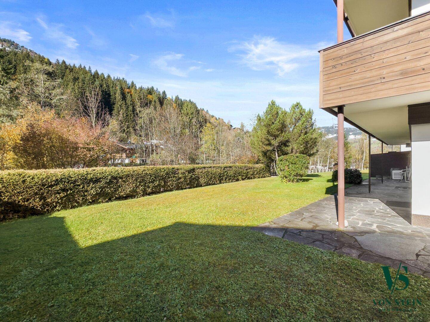 Sonniger Gartenbereich mit gepflegtem Rasen, Steinterrasse und Blick auf bewaldete Berge.