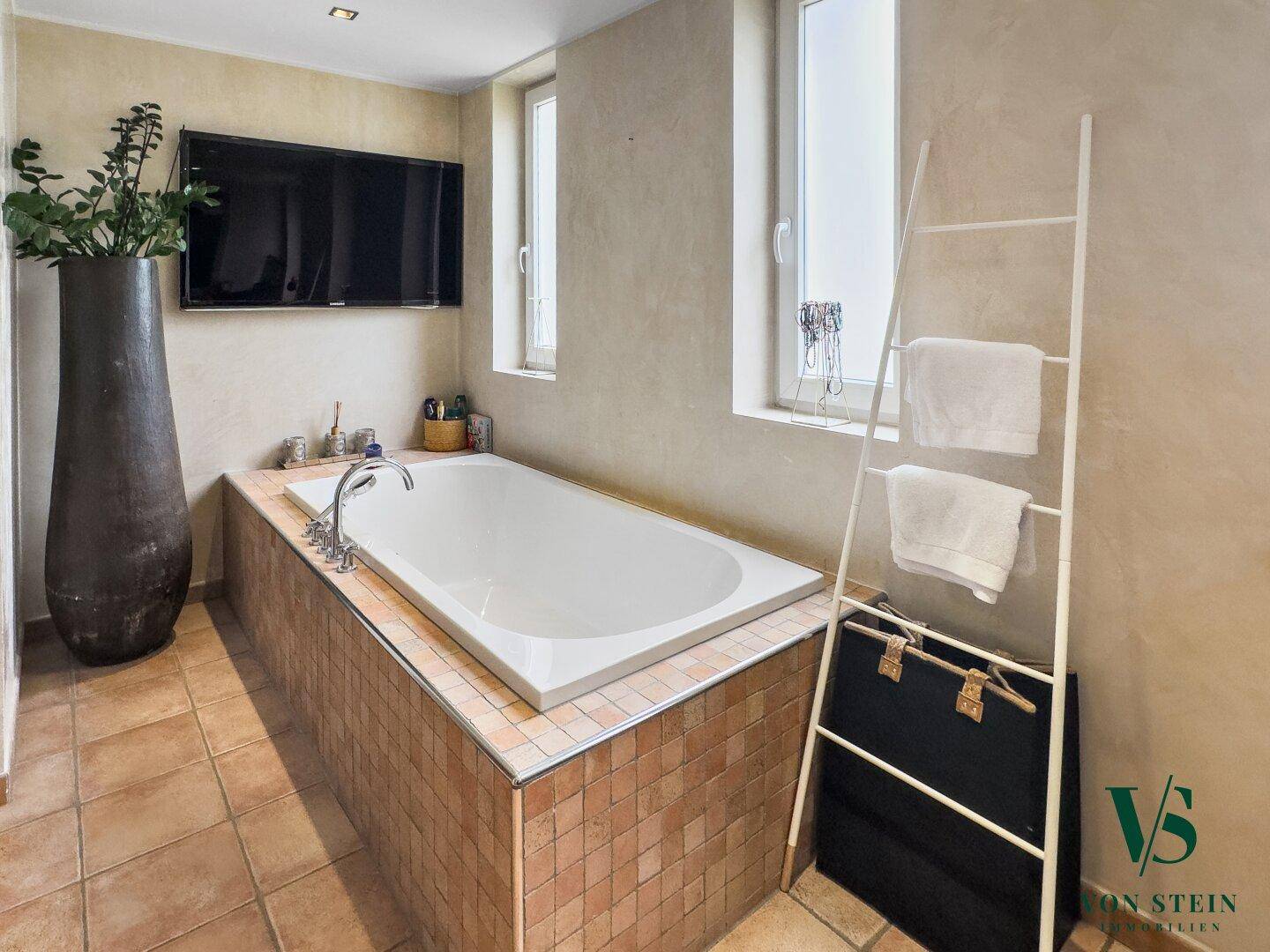 Elegante Badewanne in gefliester Umrandung mit Flachbildfernseher und Fenster.