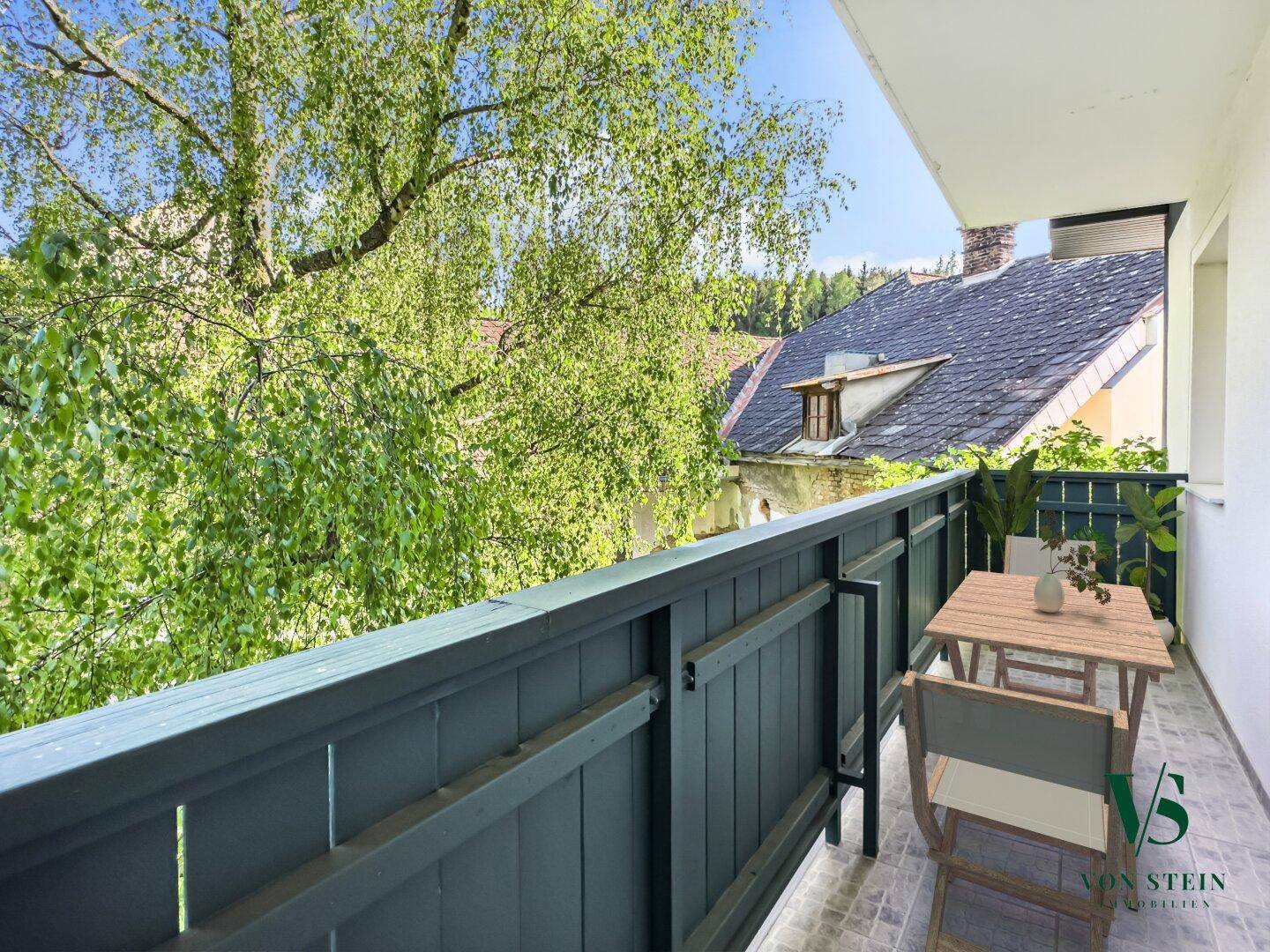 Kleiner Balkon mit Tisch und Stühlen, umgeben von üppigem Grün und Blick auf Dächer.
