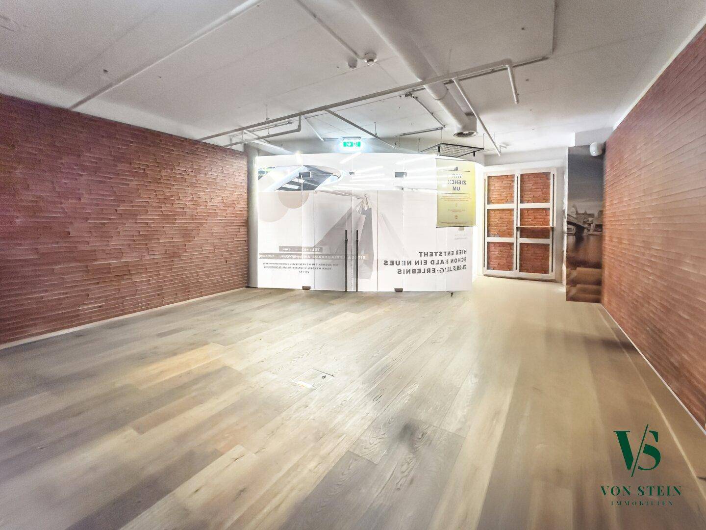 Großzügiger Gewerberaum mit Ziegelwand, Holzboden und einer modernen, halbtransparenten Trennwand.