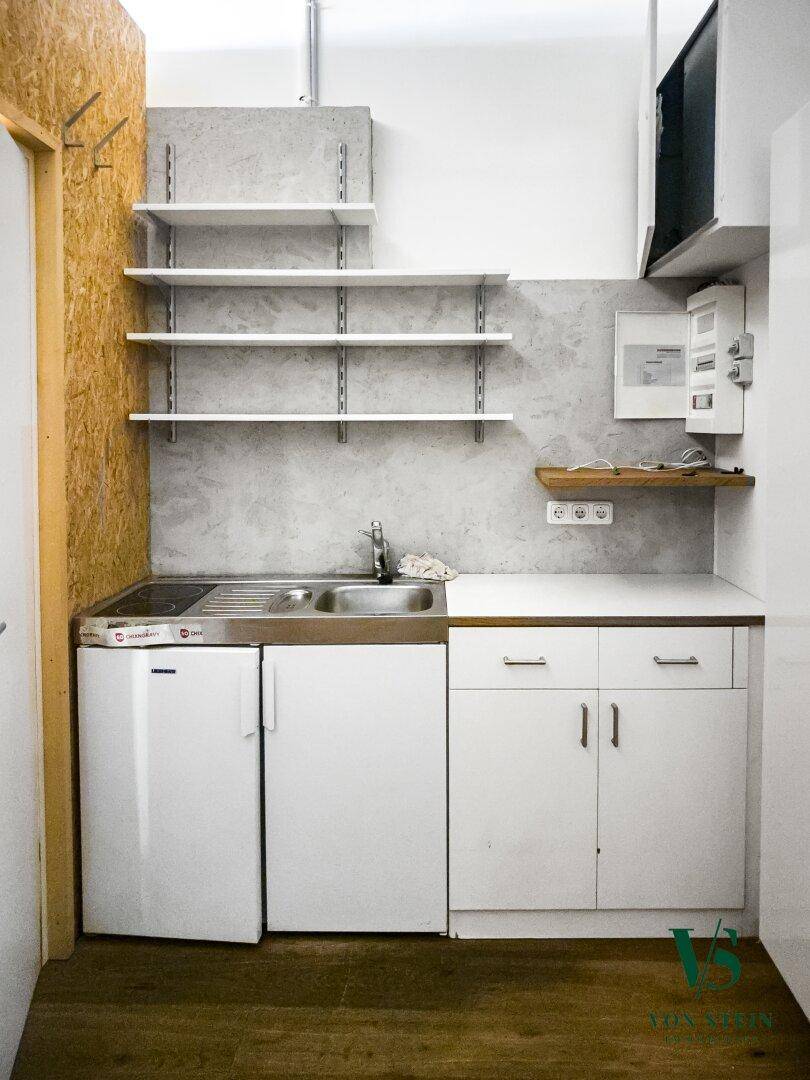 Kleine Küchenzeile mit Spüle, Kochfeld, Kühlschrank und offenen Regalen in einem kompakten Raum.