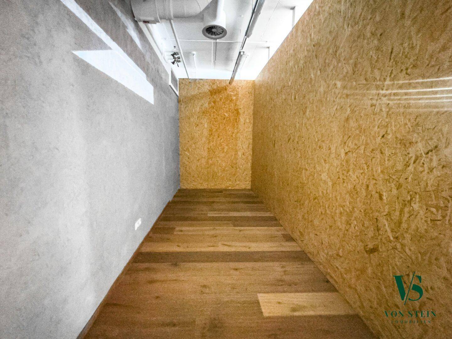 Schmaler Raum mit OSB-Plattenwänden und hellem Holzboden, der als Durchgang oder Lager dienen könnte.