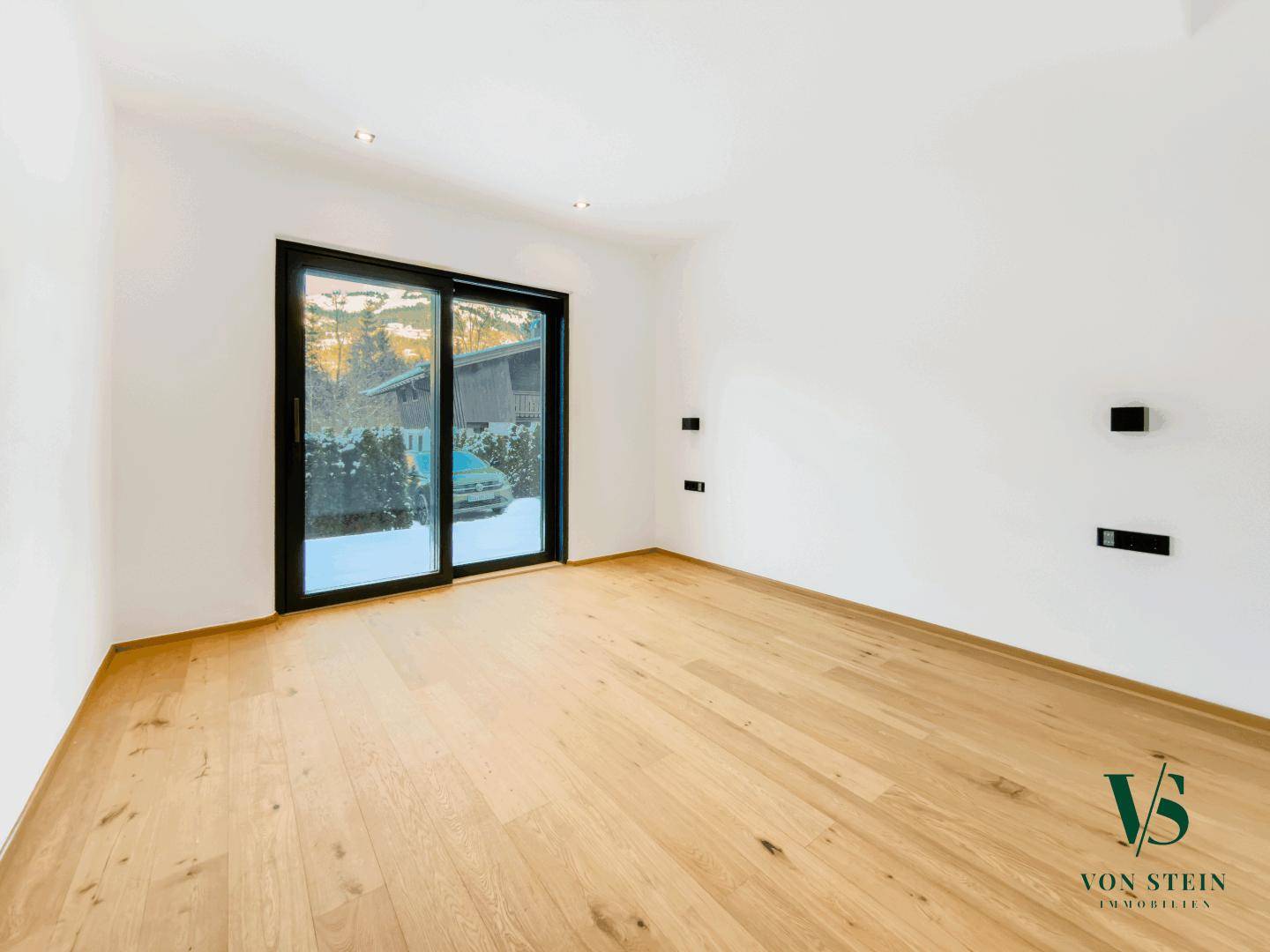 Leeres Zimmer mit weiß gestrichenen Wänden, Holzboden und Schiebetür zum Außenbereich.