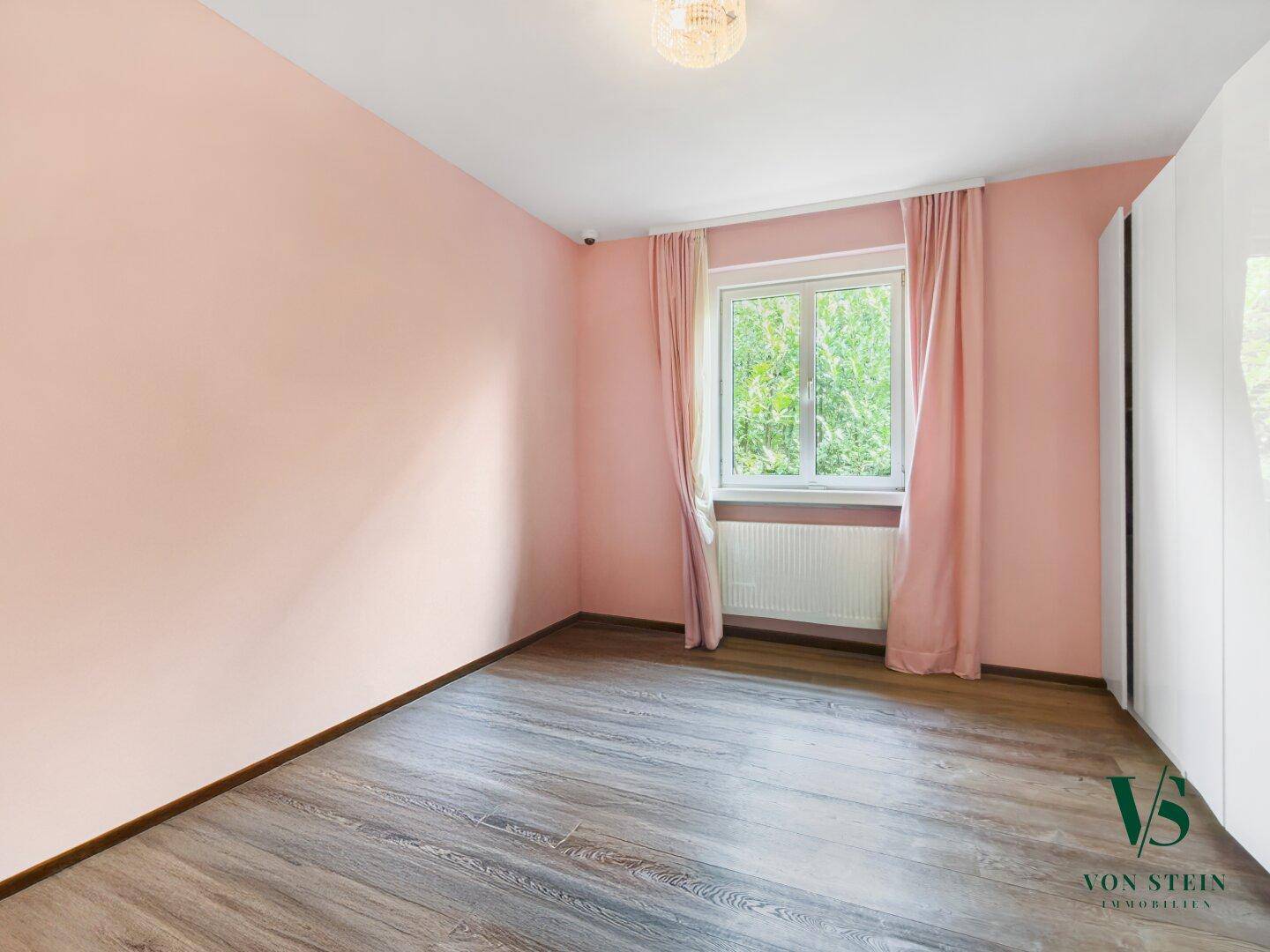 Gemütliches Schlafzimmer mit rosafarbenen Wänden, Laminatboden und einem Fenster mit Blick ins Grüne.