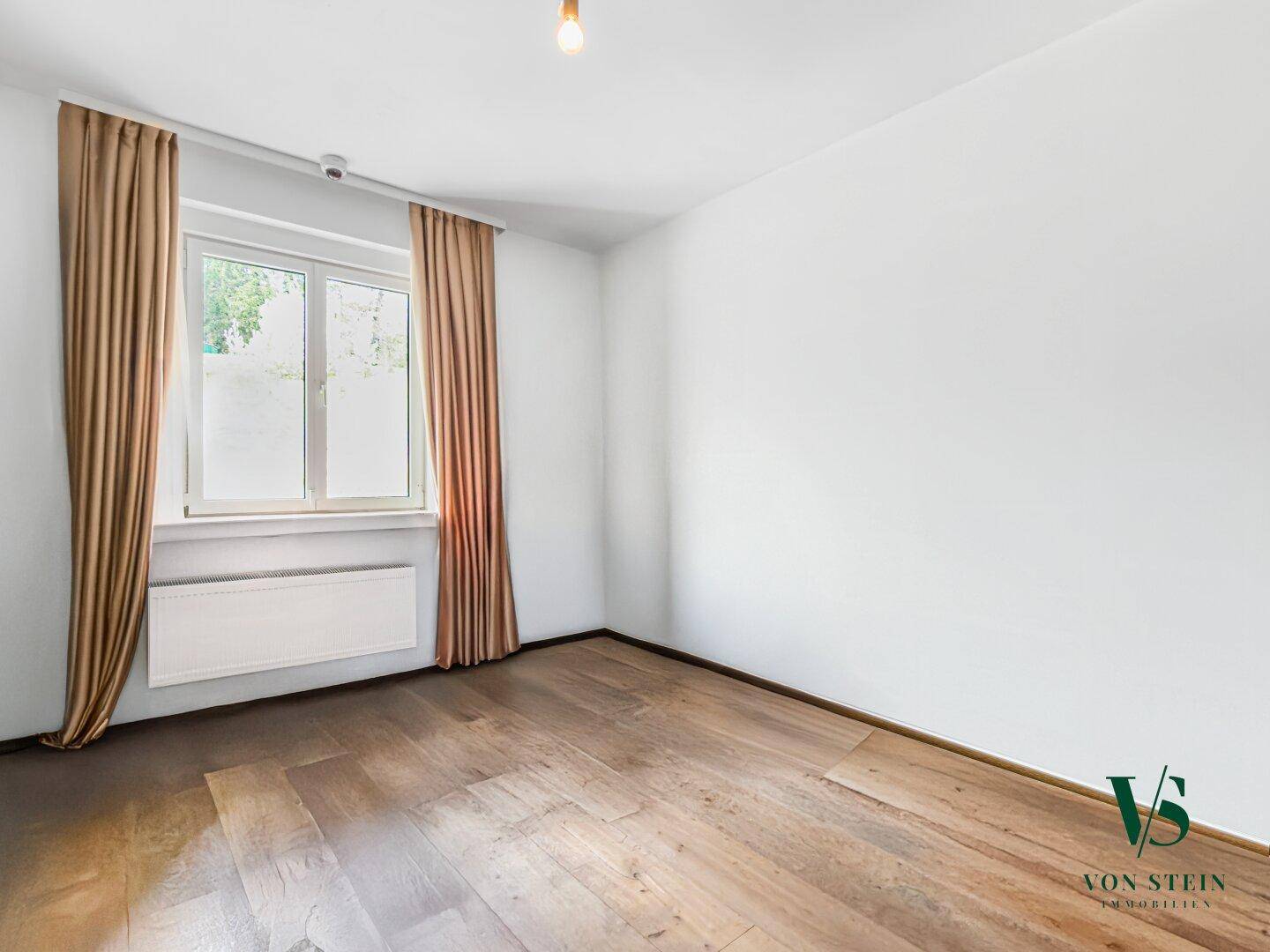Helles Zimmer mit weißen Wänden, Parkettboden und einem Fenster mit braunen Vorhängen.