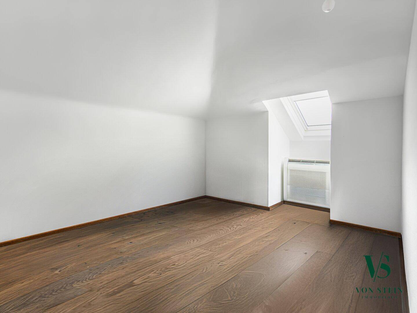 Helles Dachzimmer mit schrägen Wänden, Dachfenster und dunklem Parkettboden.