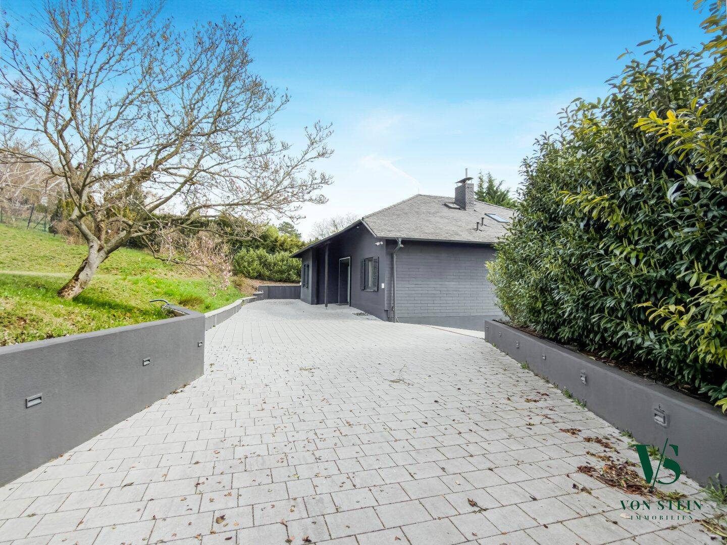 Zufahrt zum Haus mit gepflastertem Weg, umgeben von gepflegten Grünflächen und Bäumen.