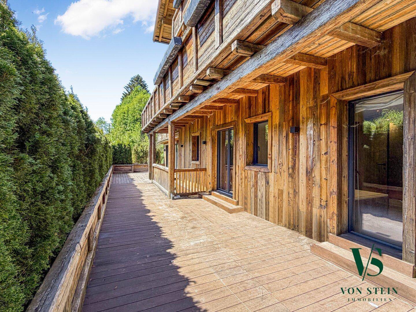 Lange Holzterrasse entlang des Hauses mit grüner Hecke und Zugang zu den Innenräumen.
