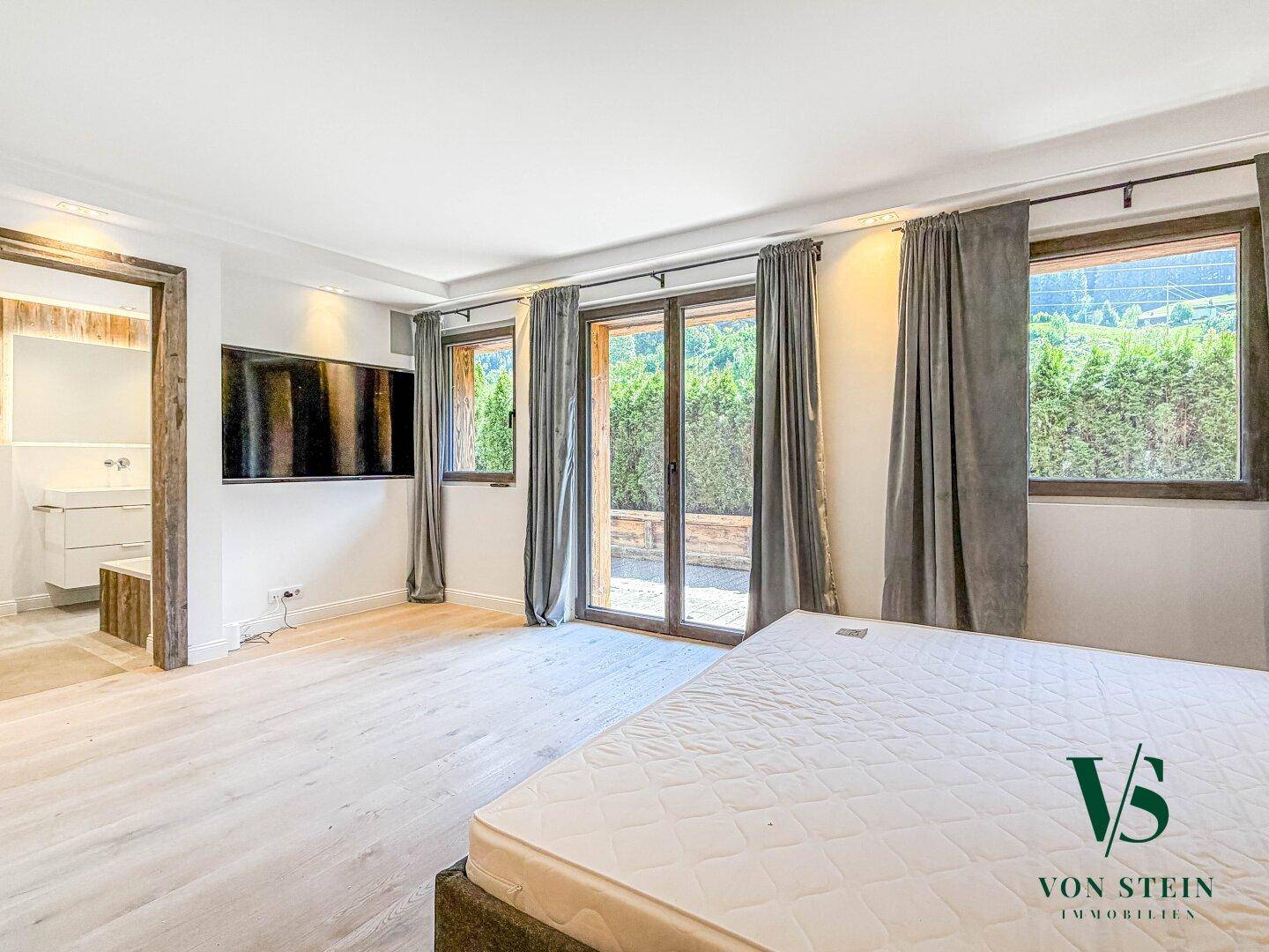 Geräumiges Schlafzimmer mit Holzboden, großen Fenstern und direktem Zugang zum Bad.