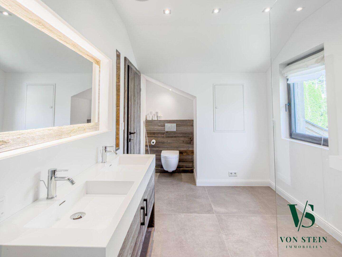 Helles Badezimmer mit Doppelwaschbecken, Toilette und Fenster unter Dachschräge.