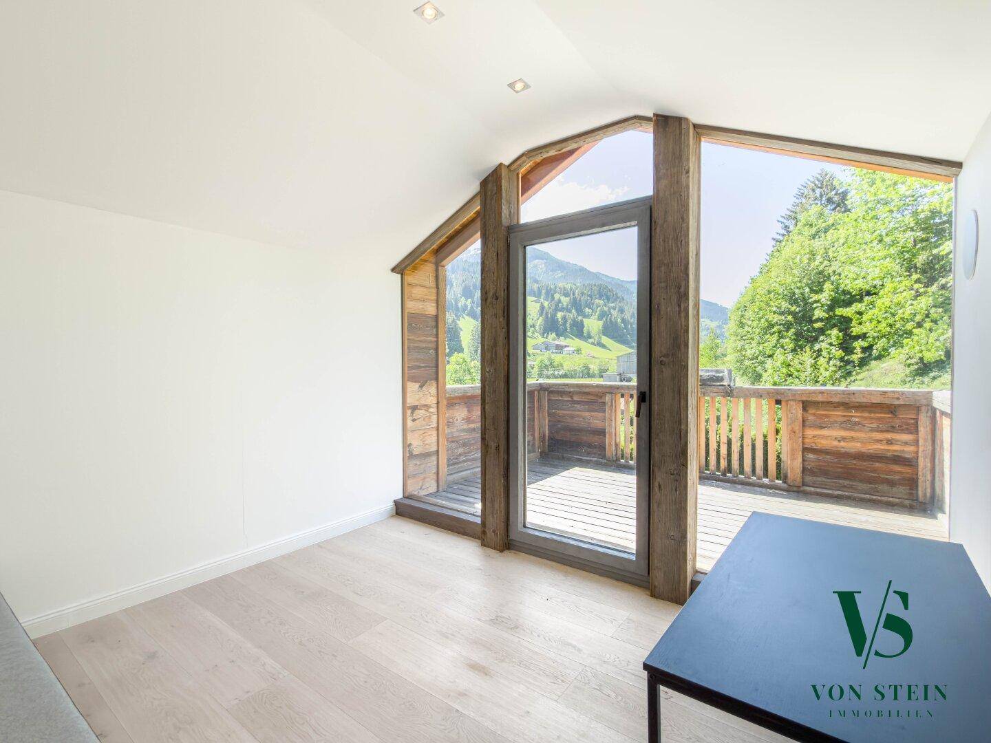 Helles Zimmer mit Holzboden, großen Fenstern und Zugang zu einem Balkon mit Bergblick.
