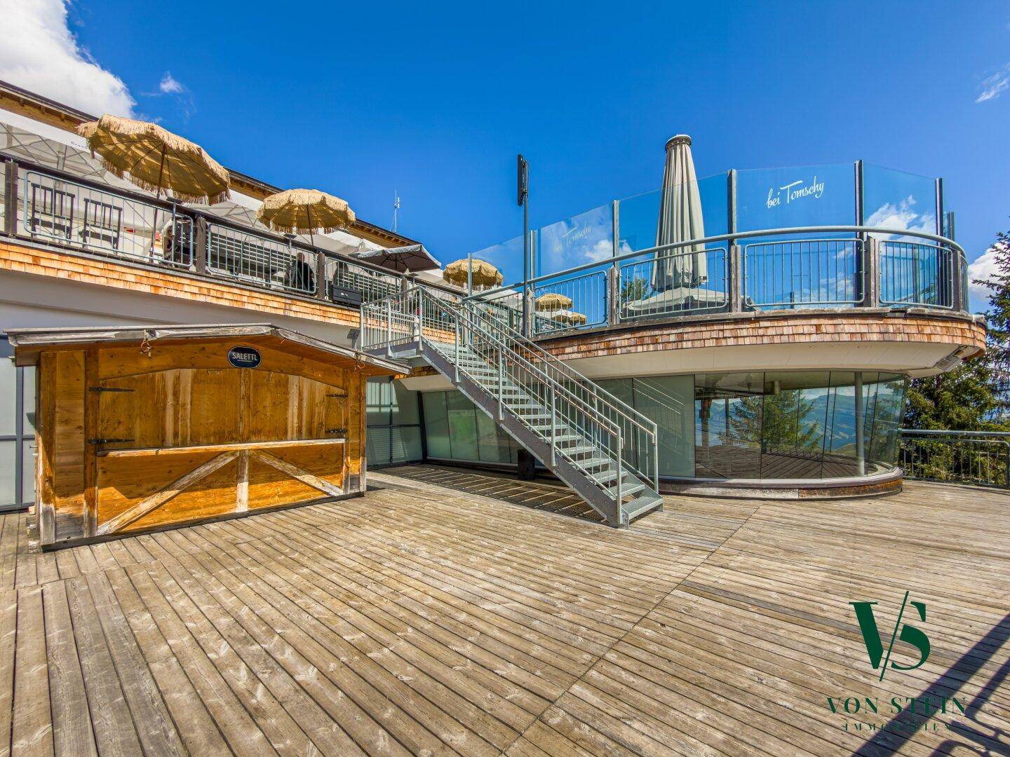 Geräumige Holzterrasse mit Metalltreppe und Glasgeländer, umgeben von einem modernen Gebäude mit Panoramablick.