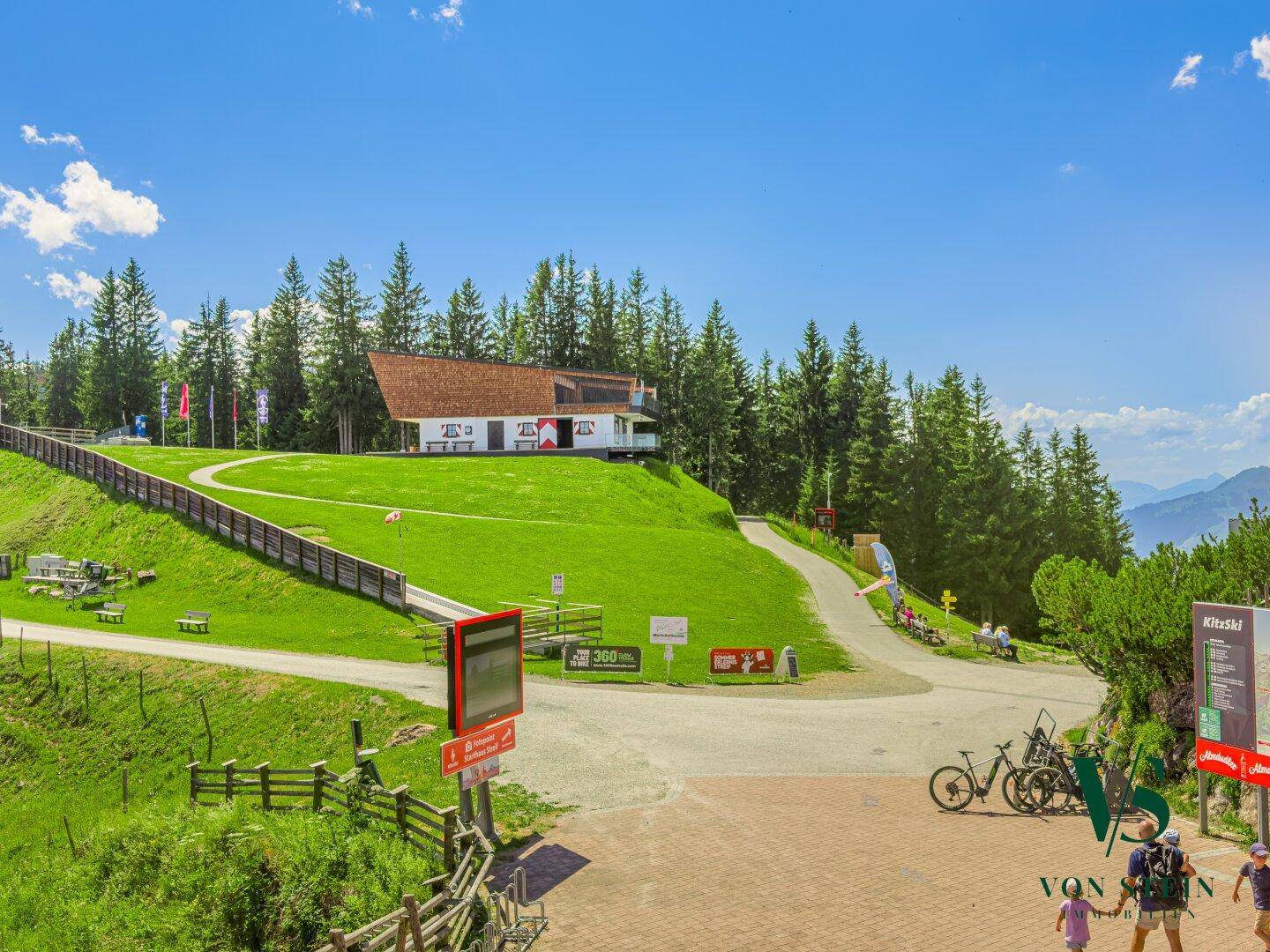Weitläufige grüne Berglandschaft mit einem Gebäude im Hintergrund, umgeben von Bäumen unter blauem Himmel.