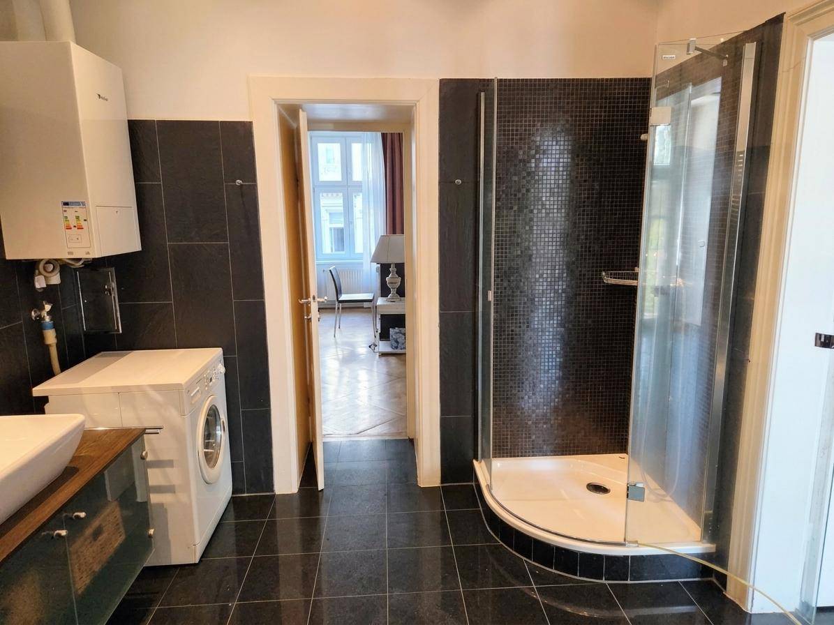 Badezimmer mit Dusche, Waschmaschine und dunklen Fliesen, mit Zugang zu einem anderen Raum.