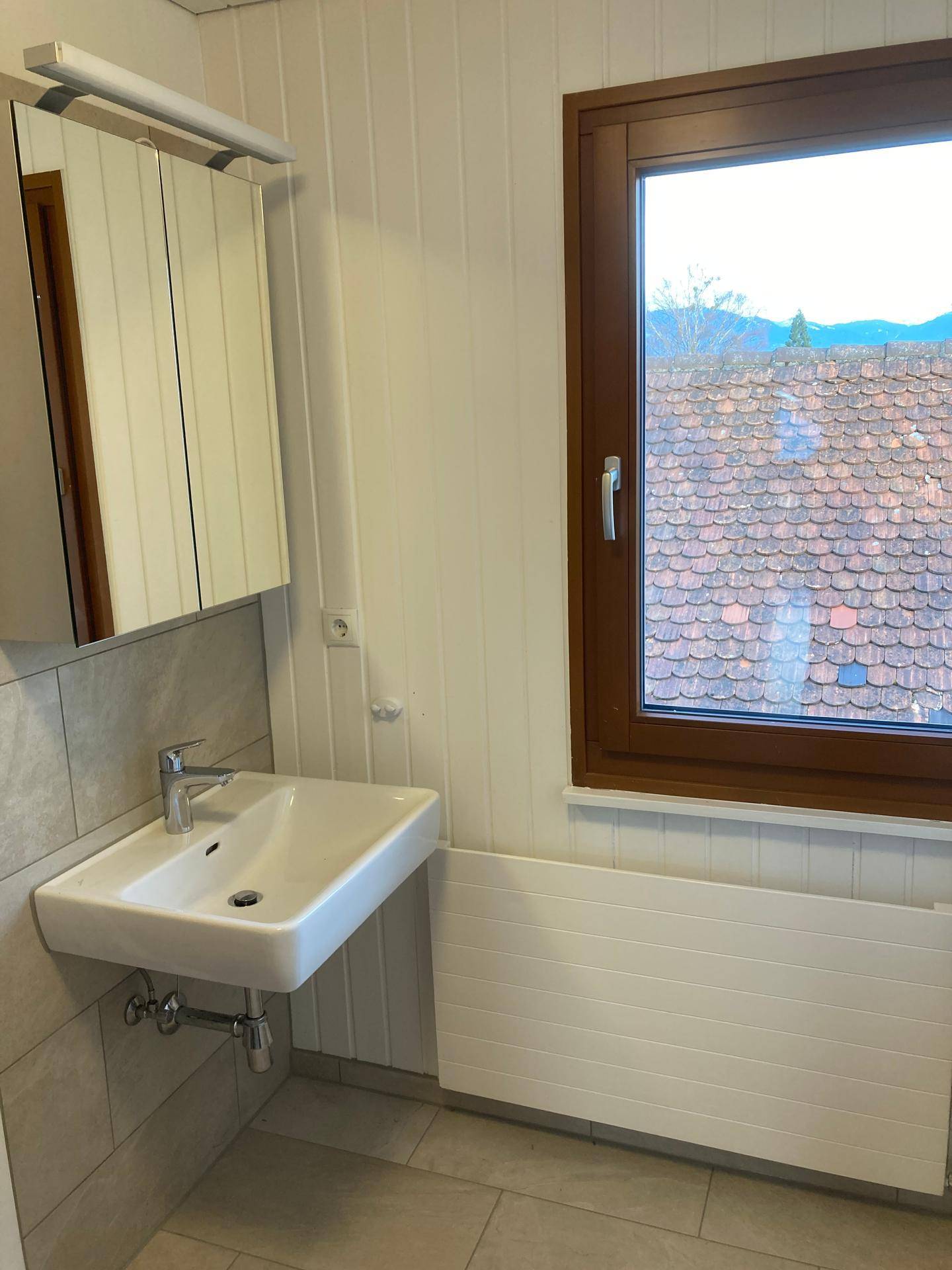 Modernes Badezimmer mit Waschbecken, Spiegelschrank, Heizkörper und einem Fenster für natürliche Belüftung.