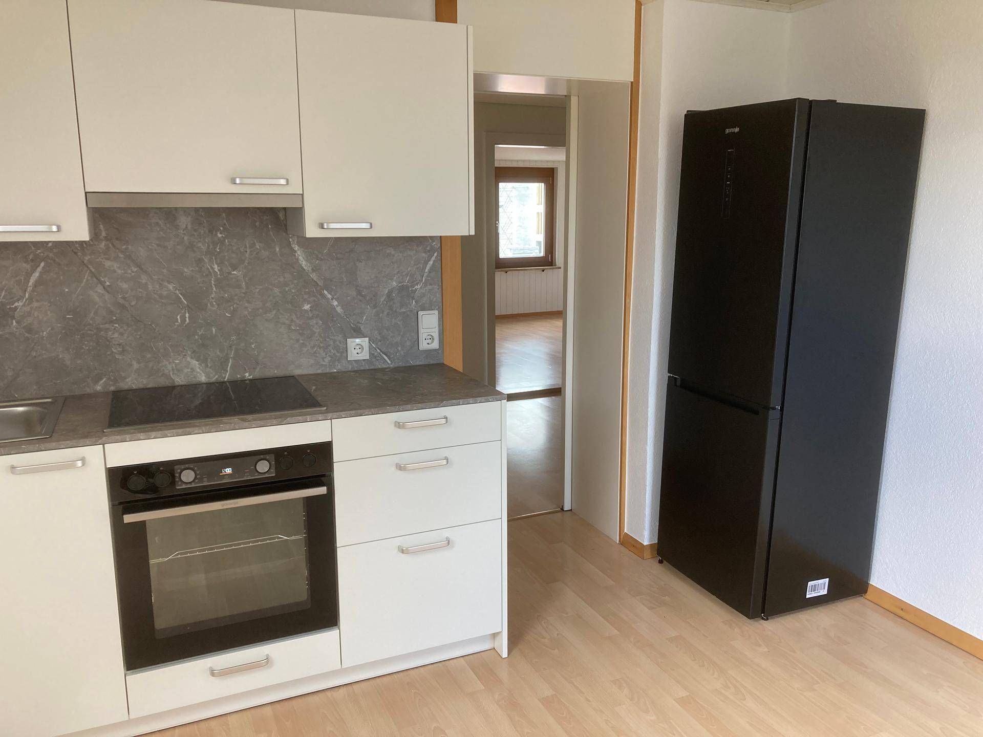 Funktionale Küche mit weißen Schränken, grauer Arbeitsplatte und einem freistehenden schwarzen Kühlschrank.