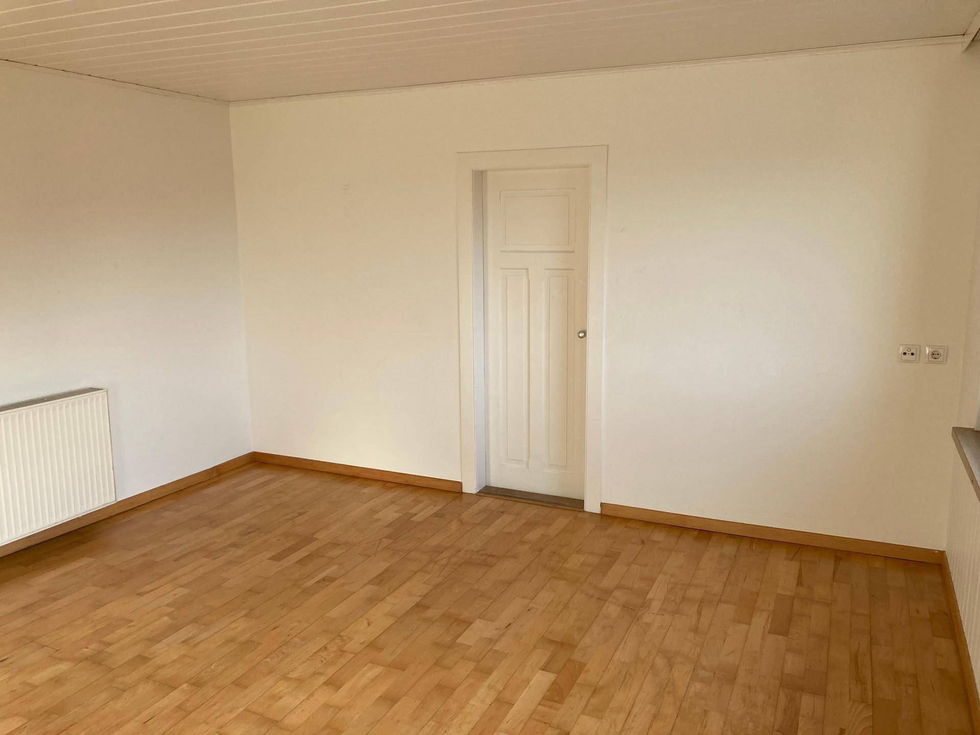 Geräumiger, leerer Raum mit hellem Parkettboden, weißen Wänden und einem Heizkörper.