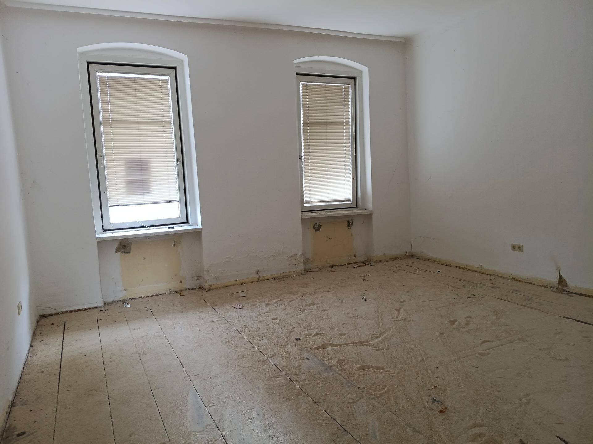 Leerer Raum mit zwei Fenstern und sichtbaren Spuren früherer Renovierungsarbeiten.