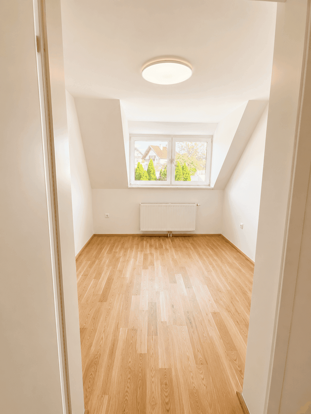 Helles Dachzimmer mit Parkettboden und zwei Fenstern, gesehen durch eine offene Tür.